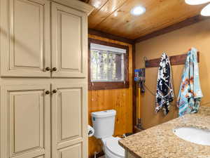 Built-in cabinetry with ample storage, a window offering natural light, wood-paneled walls, a wood-finish ceiling with recessed lighting, and a vanity with a granite-style countertop and integrated sink