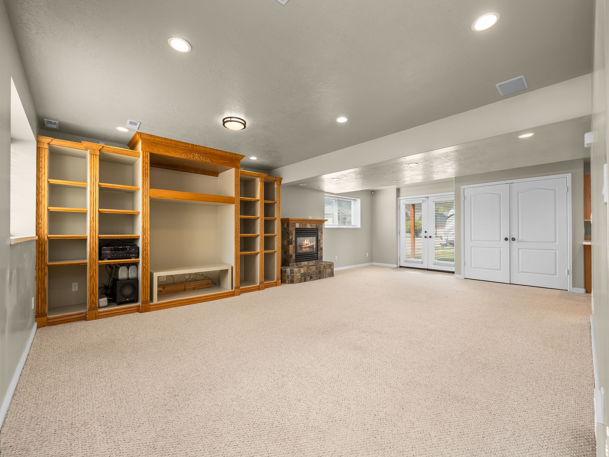 Expansive carpeted room featuring a stone-clad fireplace, built-in wood shelving, recessed lighting, and double glass-paneled doors with exterior access