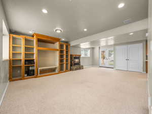 Expansive carpeted room featuring a stone-clad fireplace, built-in wood shelving, recessed lighting, and double glass-paneled doors with exterior access