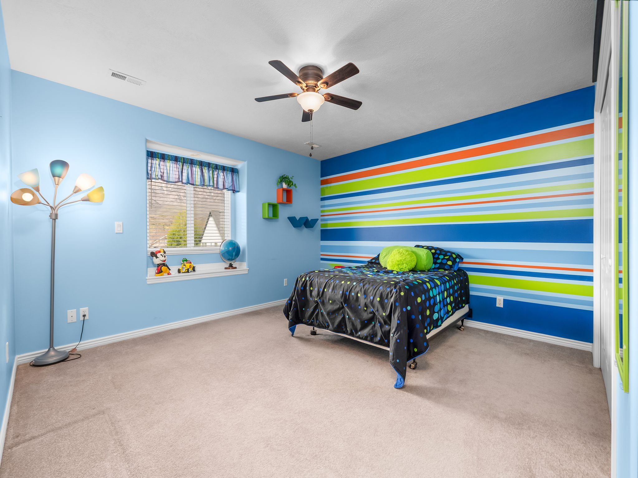 Carpeted room featuring a window with blinds, ceiling fan with integrated lighting, a colorful accent wall, and built-in shelving