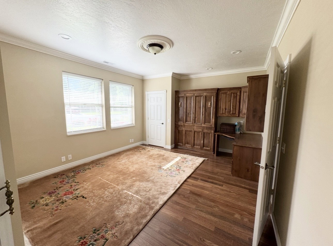 Spacious room featuring wood-finish flooring, two windows with blinds, a built-in wooden cabinetry unit, and crown molding