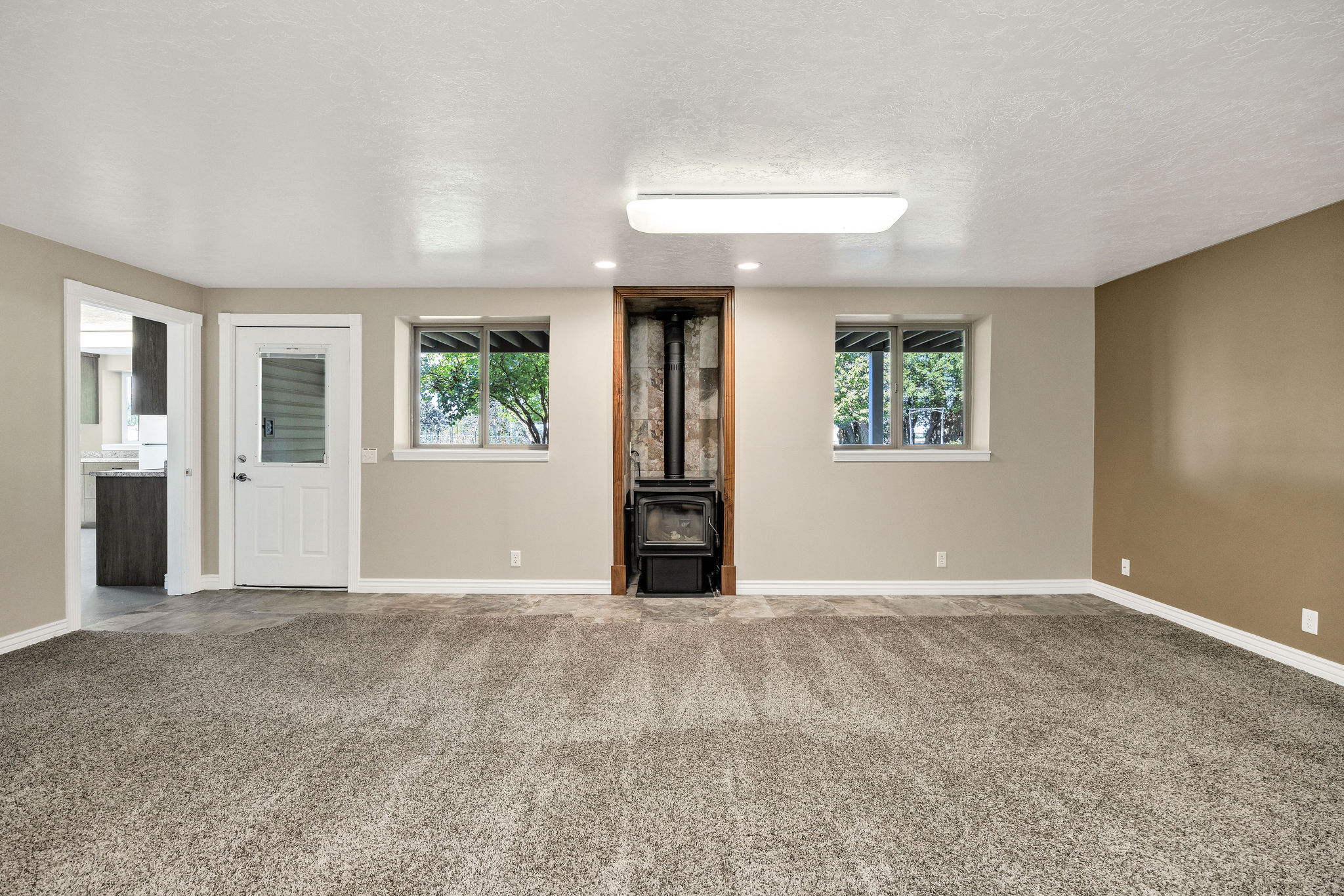 Spacious room featuring neutral paint, soft grey carpeting, a corner wood-burning stove, two windows, and a doorway leading to an additional space with wood-finish cabinetry