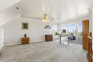 Spacious carpeted room featuring multiple windows, a ceiling fan, crown molding, and a partial view of a wood-finish staircase railing