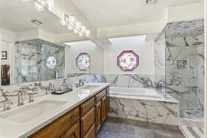 Spacious bathroom featuring a dual vanity with white countertops, a built-in soaking tub, a glass-enclosed shower, marbled wall tiling, and a decorative octagonal window