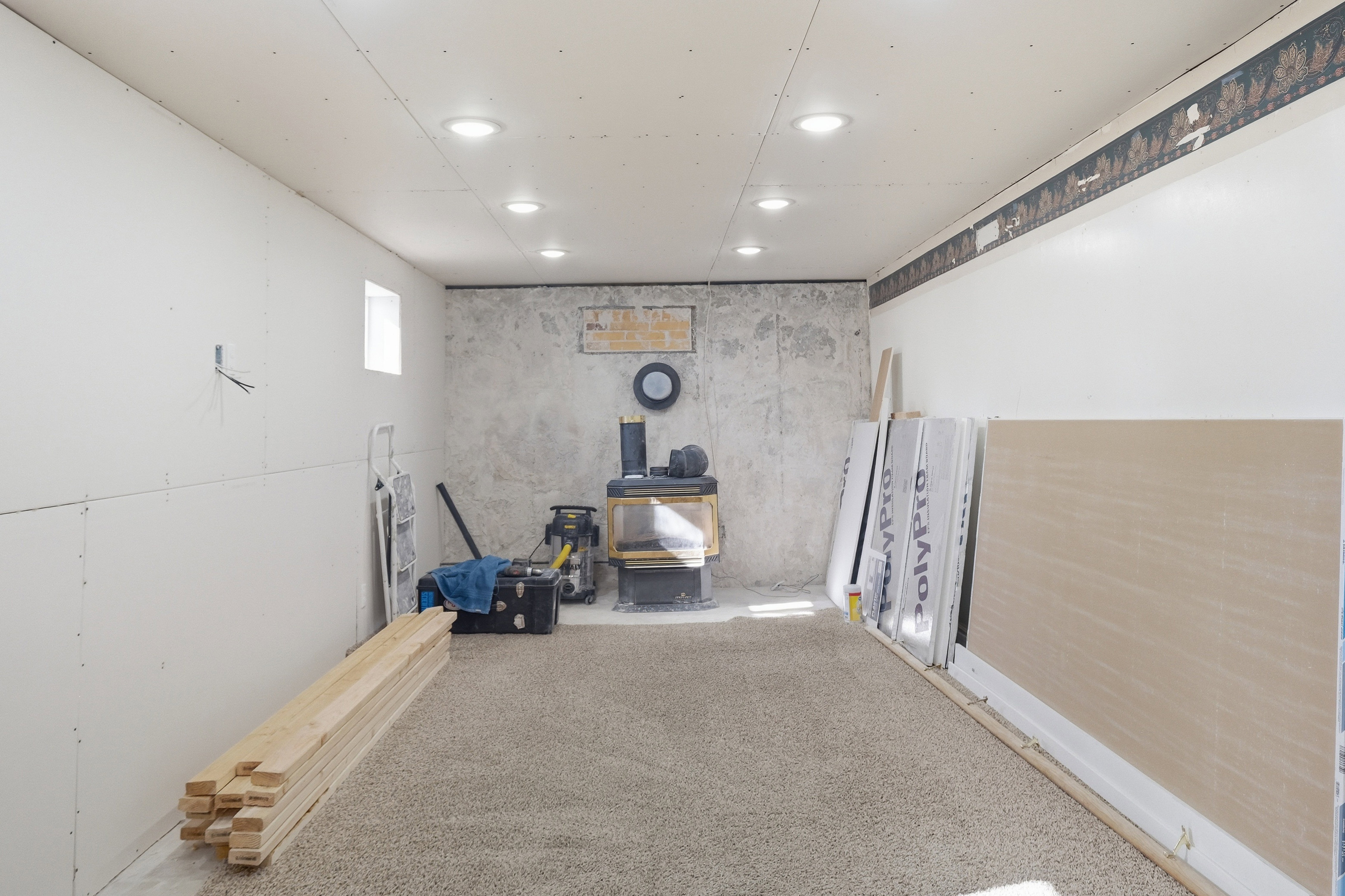 Recessed ceiling lighting illuminating a carpeted room featuring a window, partial drywall installation, and a wood-burning stove