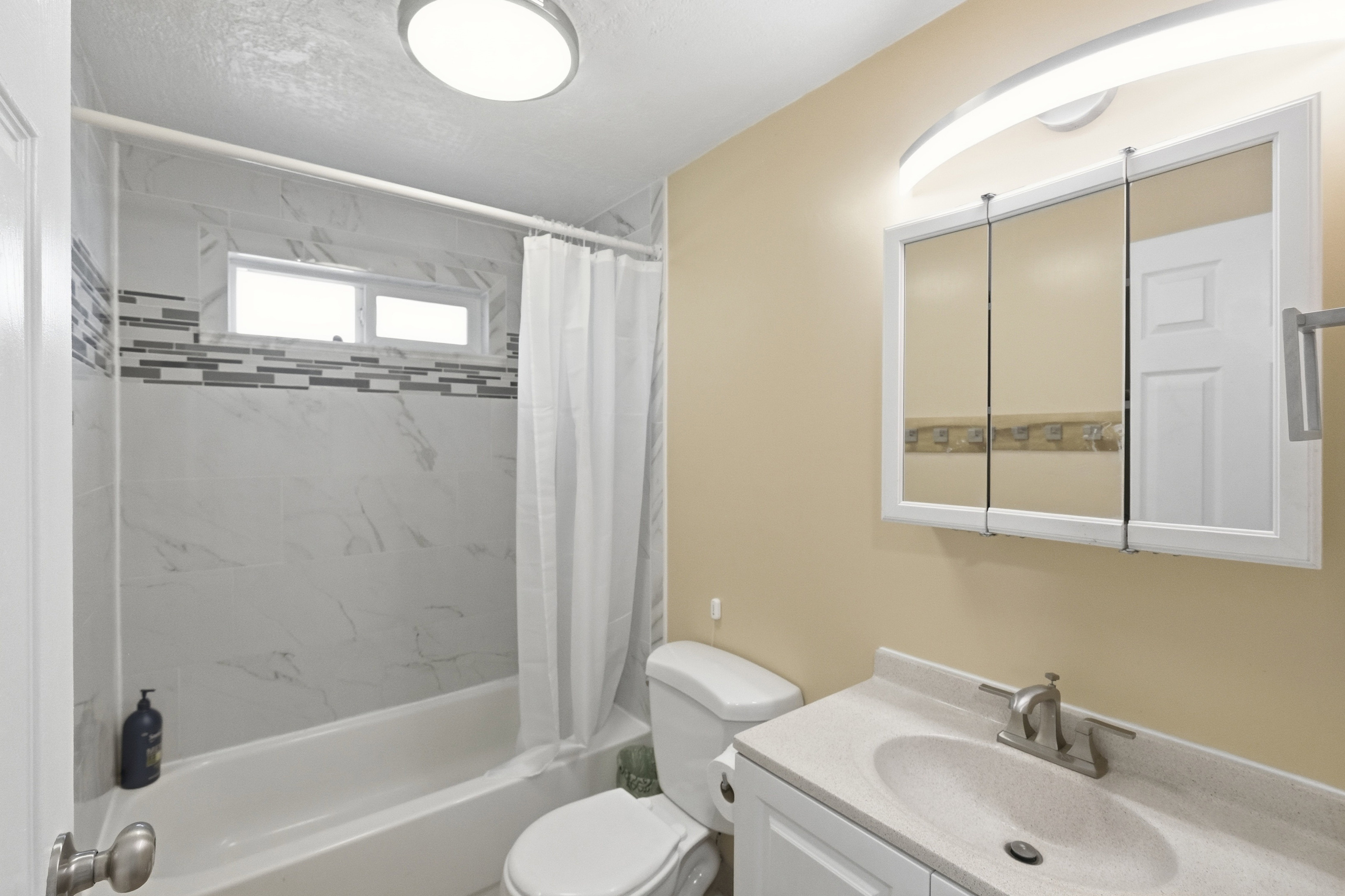 Bathroom featuring a bathtub with subway tile surround and decorative accent band, a dual-pane horizontal window, a white vanity with integrated sink, a mirrored medicine cabinet, and a wall-mounted light fixture
