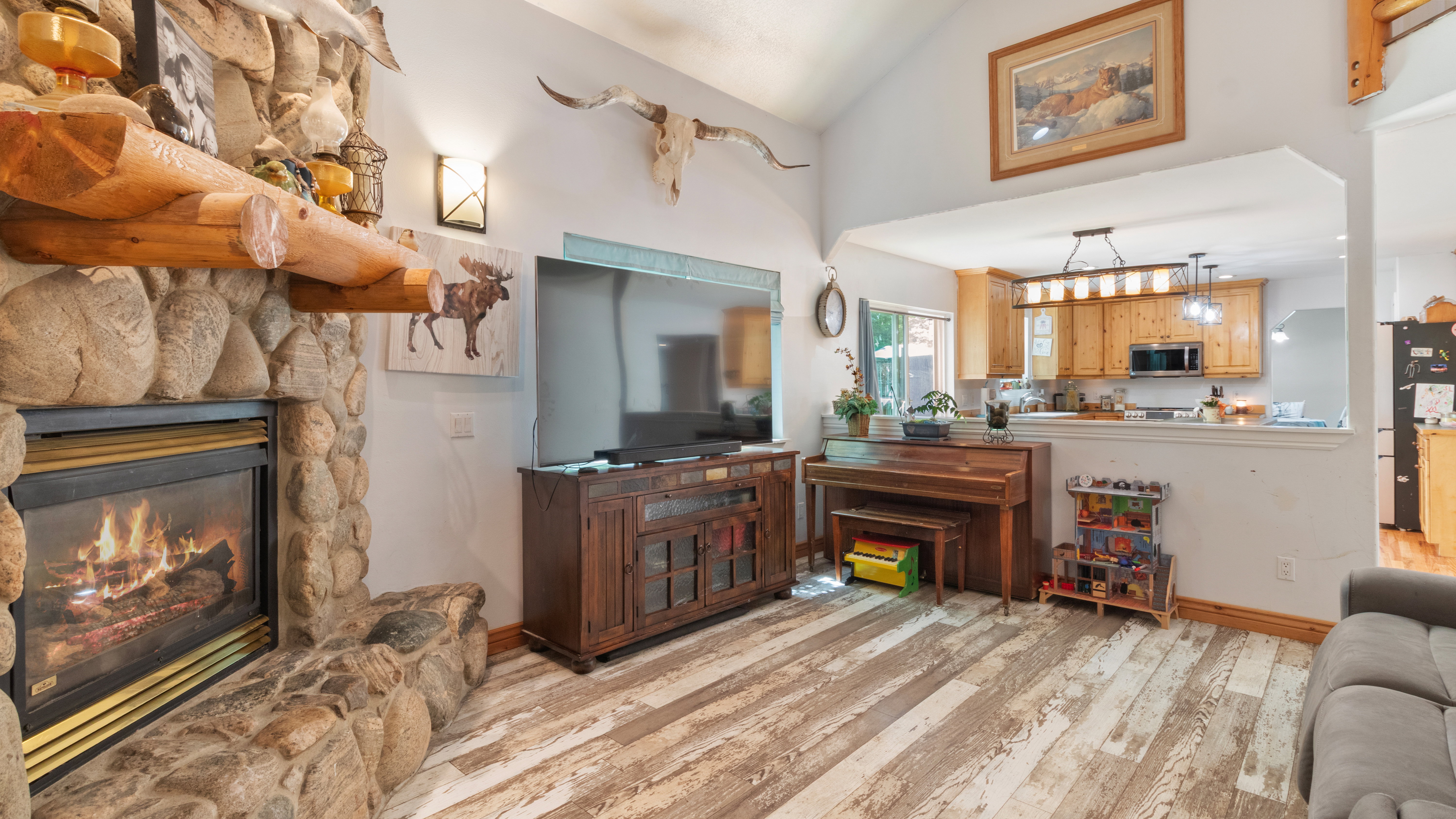 Stone-clad fireplace with a wood mantle, vaulted ceiling, wood-finish flooring, and a pass-through opening to the kitchen featuring wood cabinetry