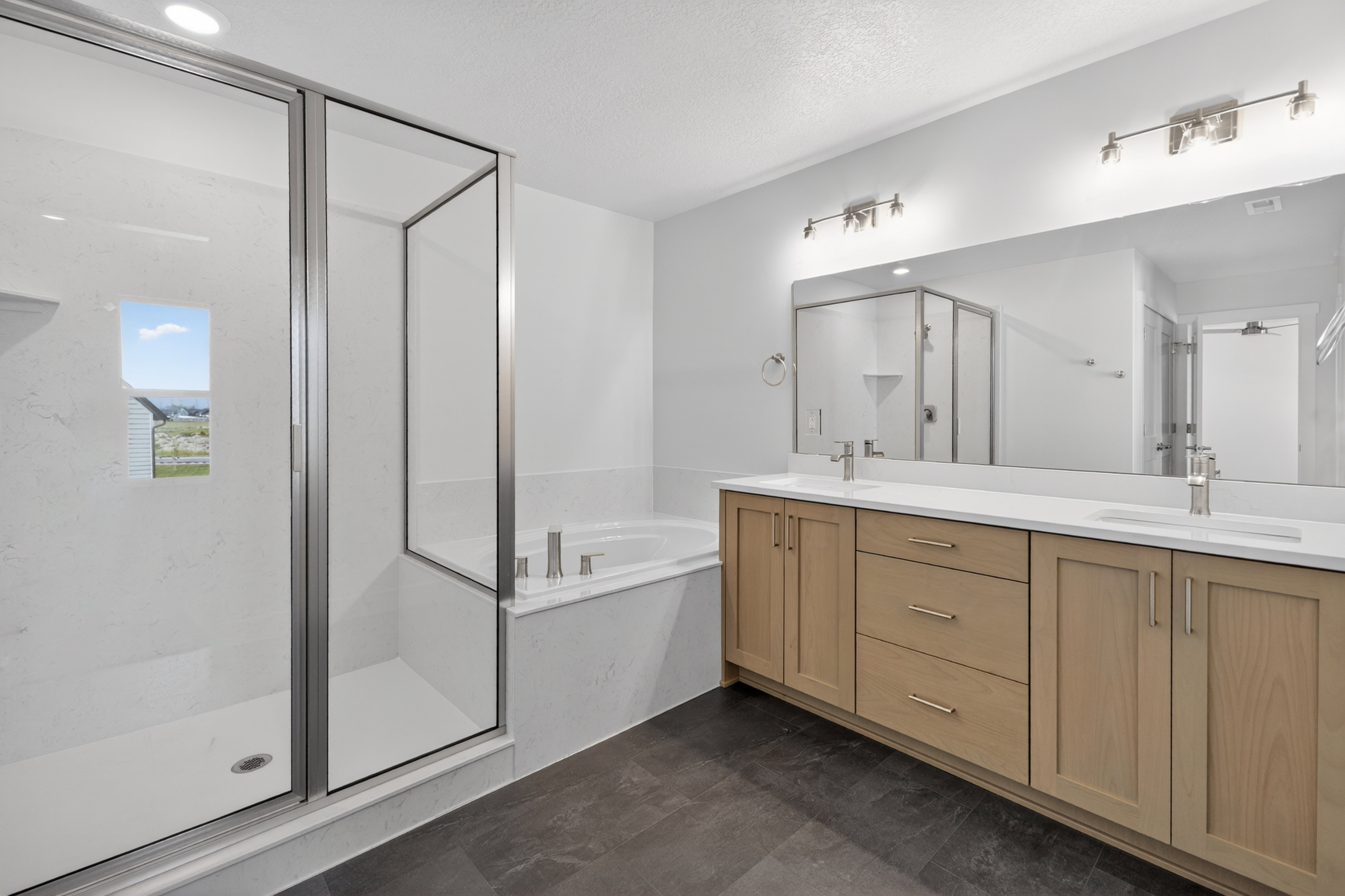 Bathroom featuring a glass-enclosed shower, a built-in soaking tub, a dual vanity with light-tone cabinetry, a large mirror, and dark-tone floor tiles
