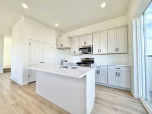 Contemporary kitchen featuring a center island with sink, light-toned cabinetry, stainless steel appliances, light countertops, and wood-finish flooring
