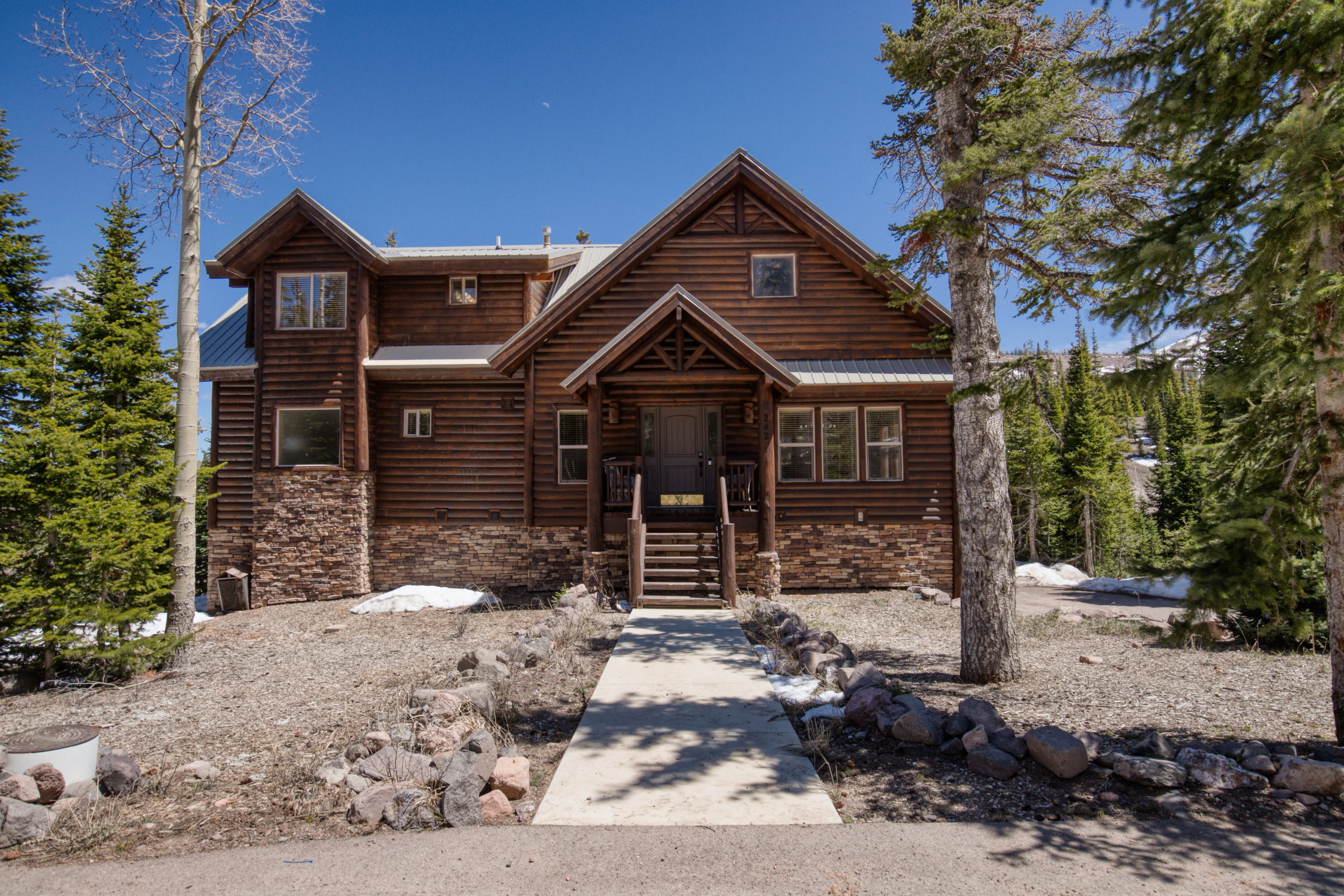Rustic cabin exterior featuring log siding, stone accents, a covered front entry, exposed wood stairs, and surrounding mature pine trees