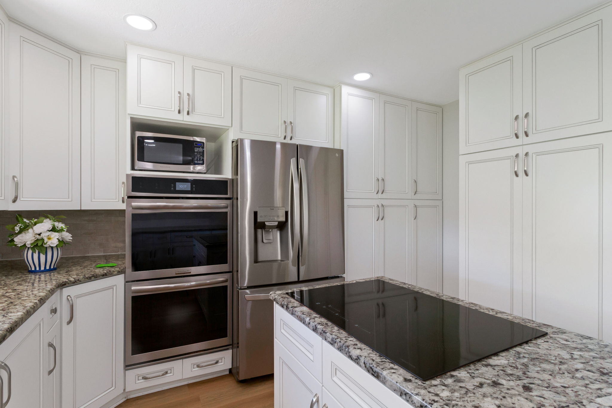 Kitchen featuring extensive white cabinetry, granite countertops, built-in double ovens, a stainless steel refrigerator, and an island cooktop