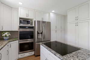 Kitchen featuring extensive white cabinetry, granite countertops, built-in double ovens, a stainless steel refrigerator, and an island cooktop