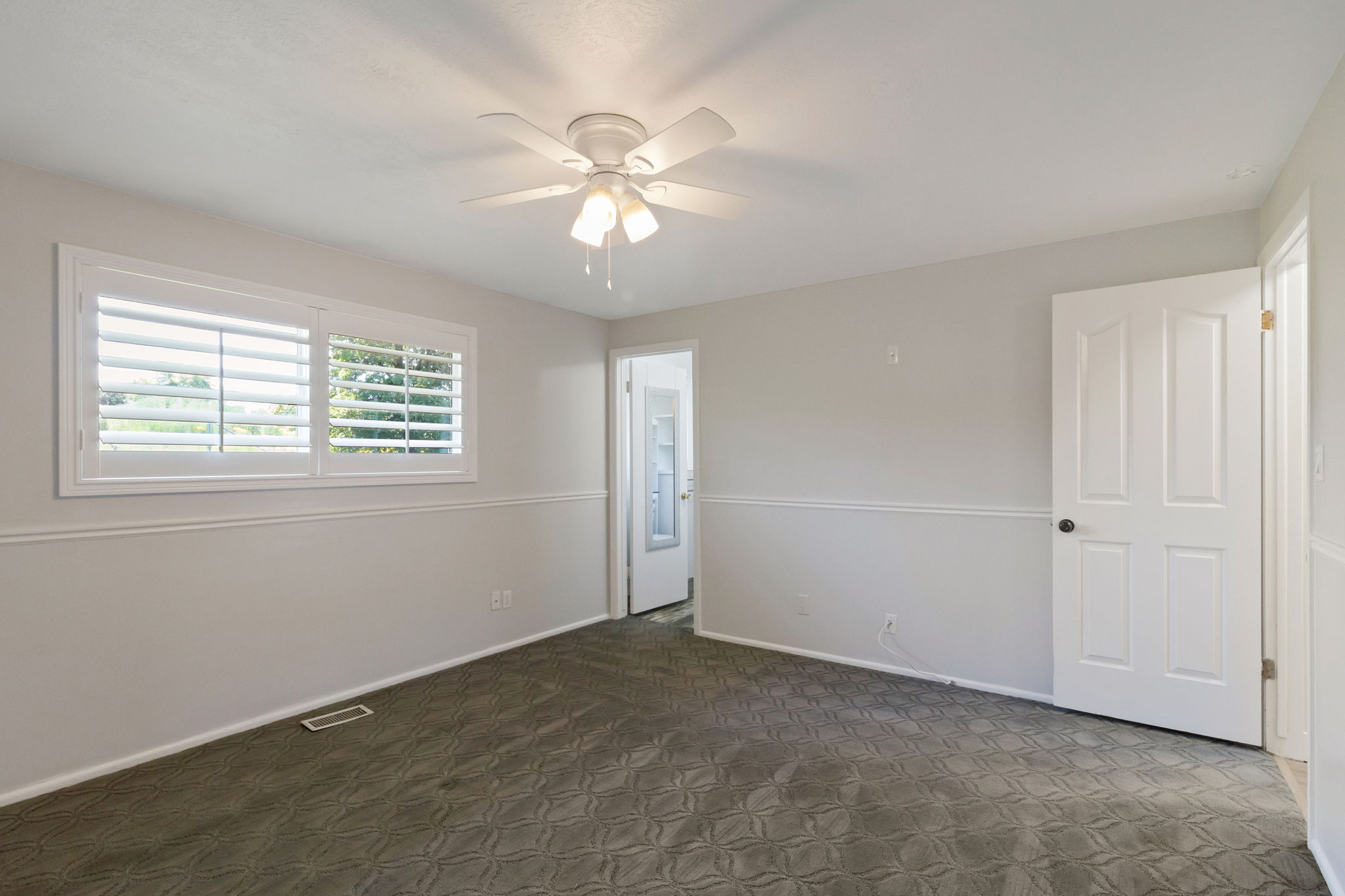 Interior room featuring a white ceiling fan with integrated lighting, window with white plantation shutters, gray patterned carpeting, and white baseboards with chair rail molding