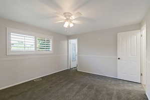 Interior room featuring a white ceiling fan with integrated lighting, window with white plantation shutters, gray patterned carpeting, and white baseboards with chair rail molding