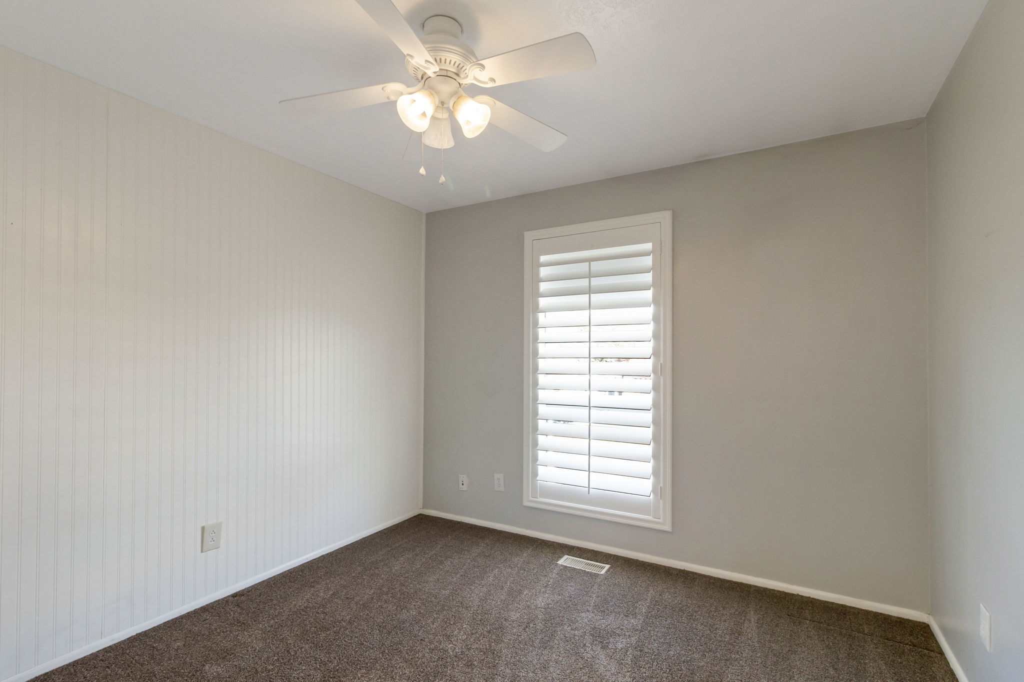 Neutral-toned room featuring a white ceiling fan with integrated lighting, a window with plantation shutters, vertical plank wall paneling, and carpet flooring