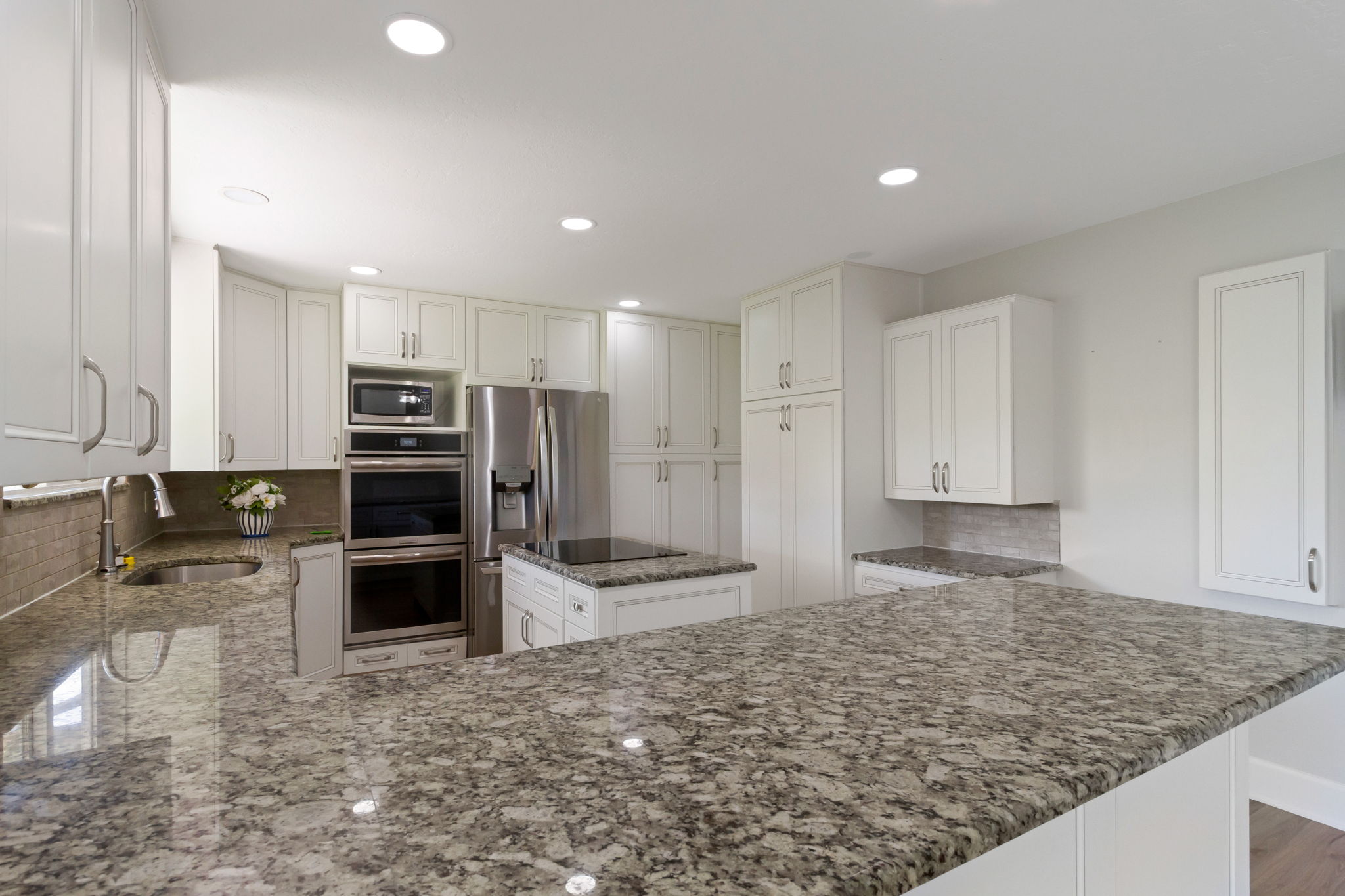 Spacious kitchen featuring extensive granite countertops, white cabinetry with brushed nickel hardware, stainless steel appliances including a built-in microwave and double ovens, and a tile backsplash