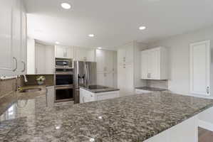 Spacious kitchen featuring extensive granite countertops, white cabinetry with brushed nickel hardware, stainless steel appliances including a built-in microwave and double ovens, and a tile backsplash