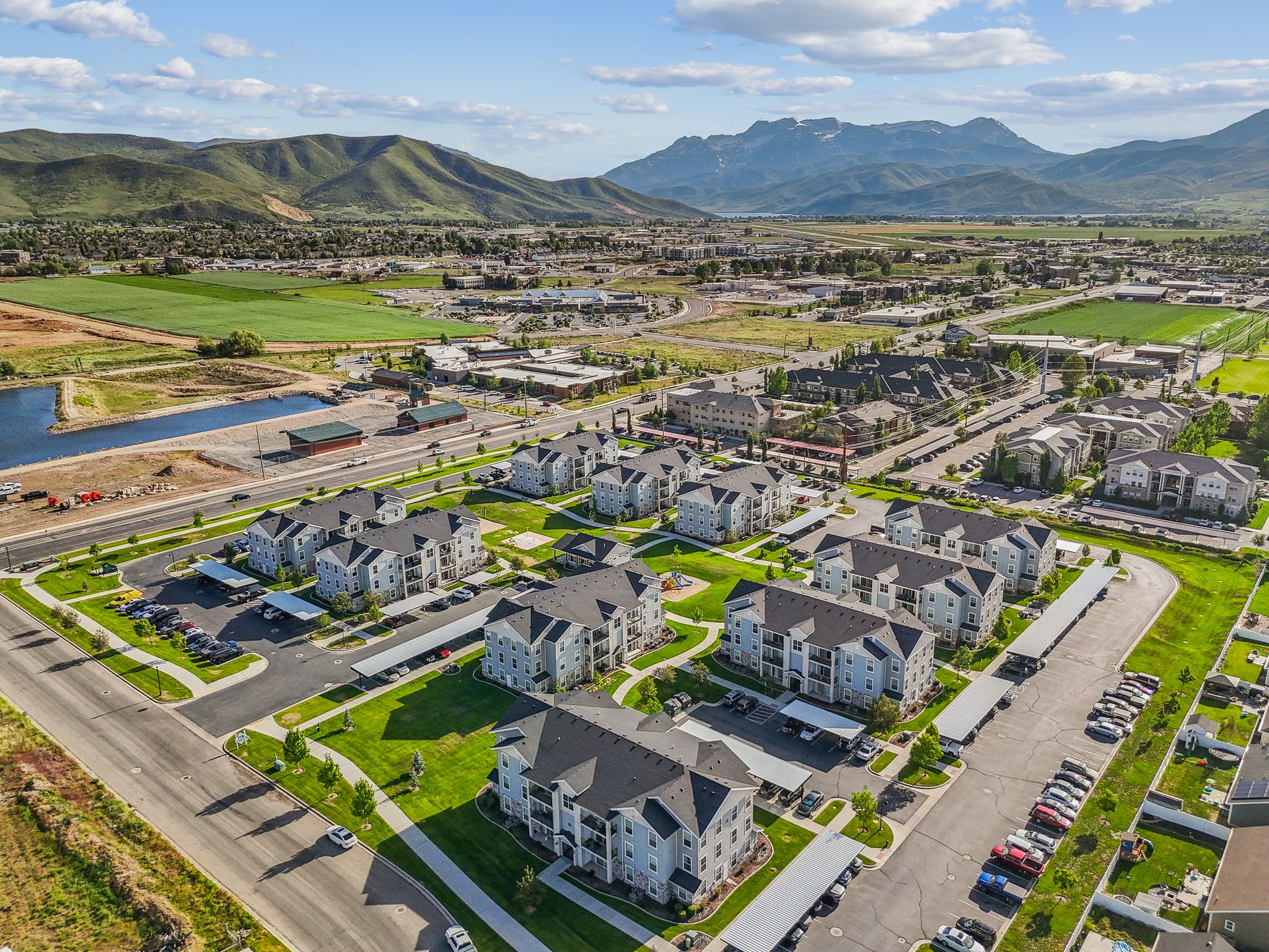 Aerial perspective of a multi-building residential community featuring covered parking structures, manicured green spaces, and a nearby pond