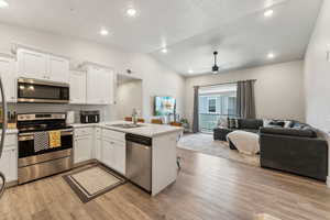 Open-concept living space featuring a kitchen with white cabinetry and stainless steel appliances, wood-finish flooring, recessed lighting, and a vaulted ceiling