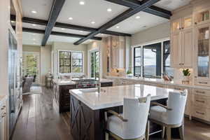 Gourmet kitchen featuring a large central island with seating, a secondary island with a professional-grade range, custom cabinetry with glass-front uppers, wood-finish flooring, and exposed ceiling beams
