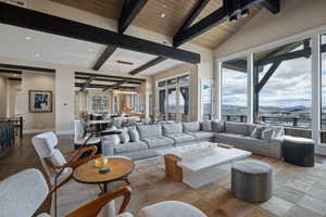 Spacious living area featuring a vaulted wood-paneled ceiling with dark exposed beams, large format floor-to-ceiling windows, light-toned wall finishes, and stone tile flooring