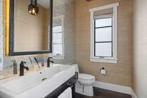 Bathroom featuring a vessel sink with dual bronze-finish faucets, a dark wood-finish vanity, a decorative framed mirror, textured wall covering, and dark wood-finish flooring