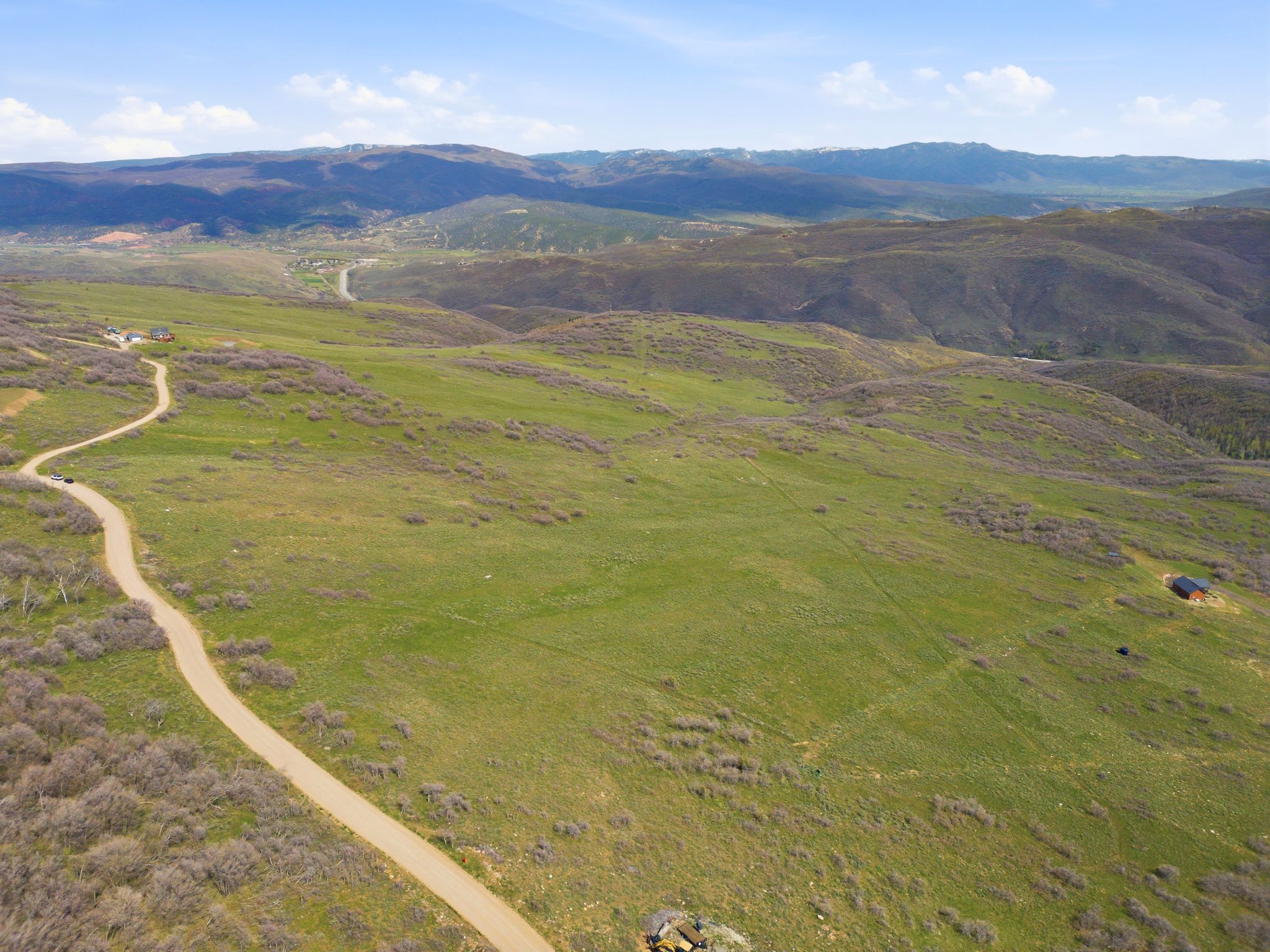 Expansive acreage featuring rolling green hills, a winding unpaved road, scattered shrubs, and distant mountain ranges under a clear sky