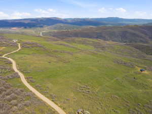 Expansive acreage featuring rolling green hills, a winding unpaved road, scattered shrubs, and distant mountain ranges under a clear sky