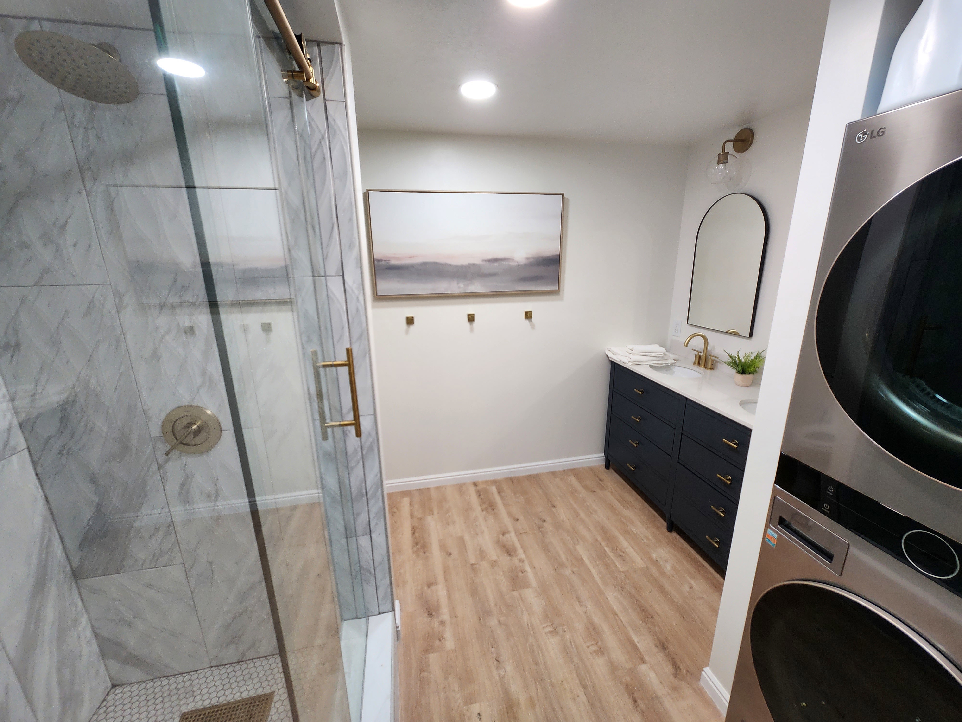 Spacious bathroom featuring a glass-enclosed shower with marble-look tile, wood-finish flooring, a long navy vanity with gold hardware, a white countertop, and a stacked washer and dryer