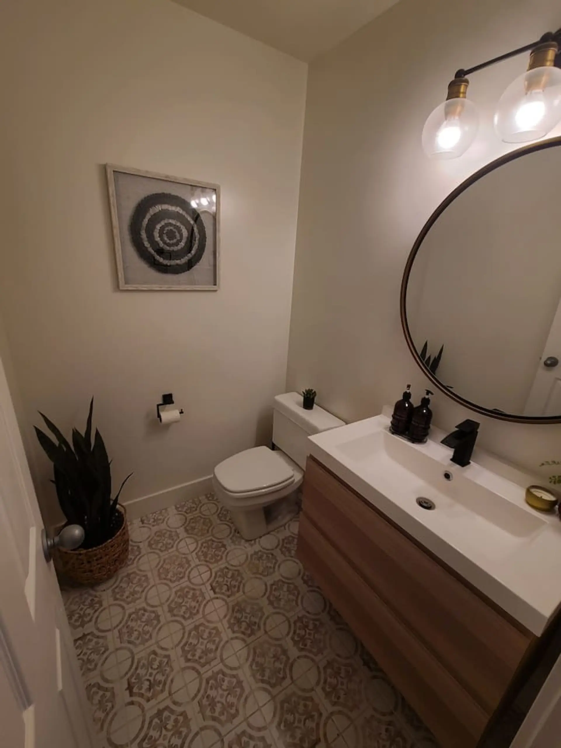 Powder room featuring patterned floor tiles, a floating wood-finish vanity with an integrated white sink, a matte black faucet, and a circular framed mirror
