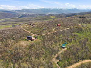 Expansive mountain landscape featuring multiple properties nestled within a wooded terrain, winding dirt roads, and distant snow-capped peaks