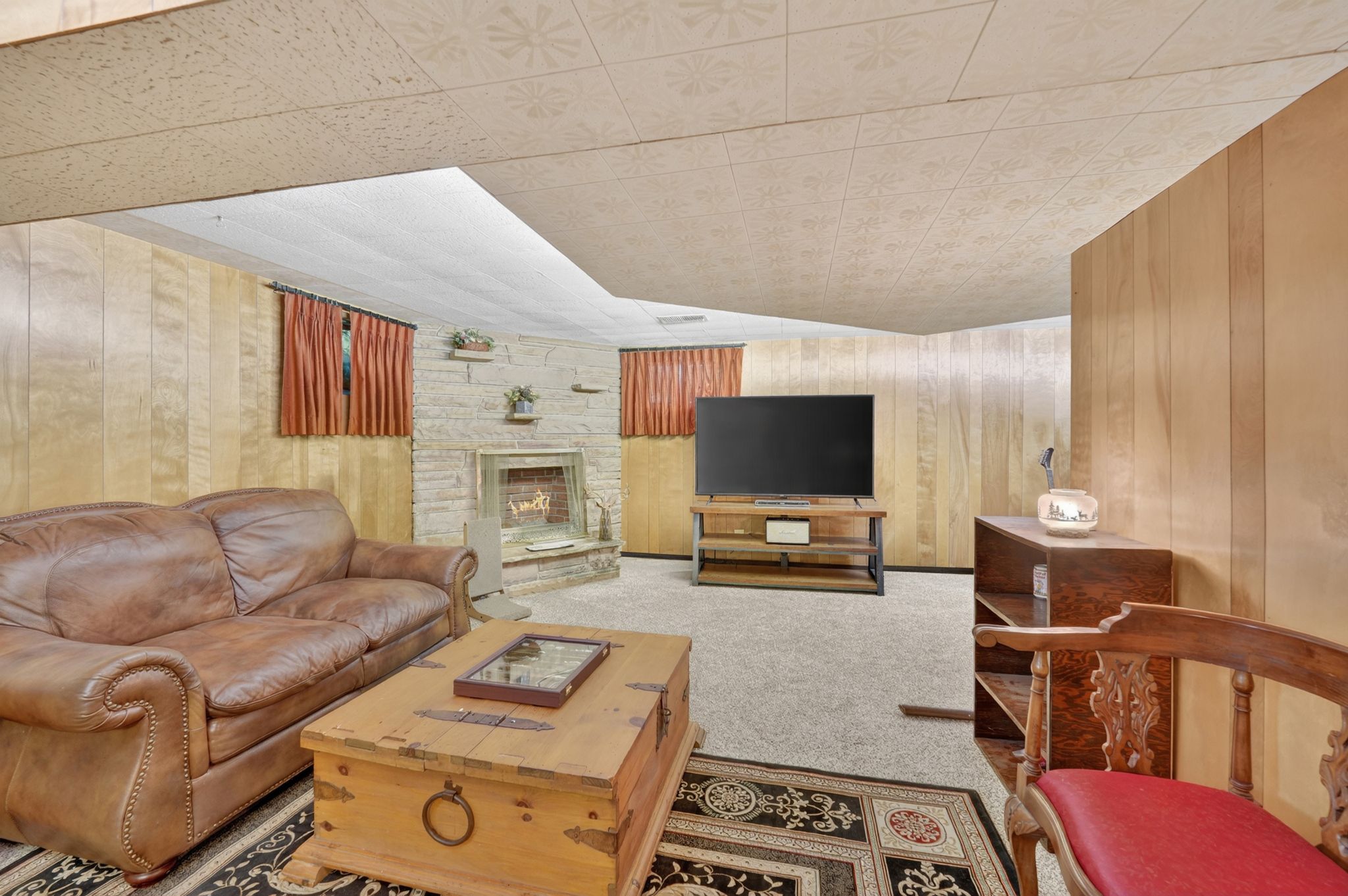 Carpeted room featuring wood paneling, a stone-faced fireplace with a hearth, and decorative ceiling tiles