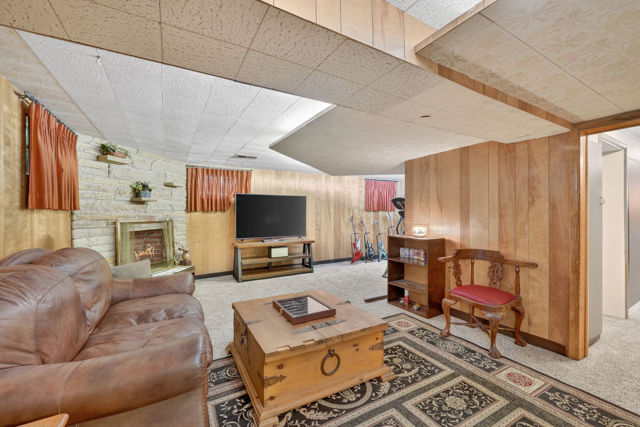 Carpeted recreation room featuring wood paneling, a stone-clad fireplace with a brass-colored firebox, and a drop ceiling