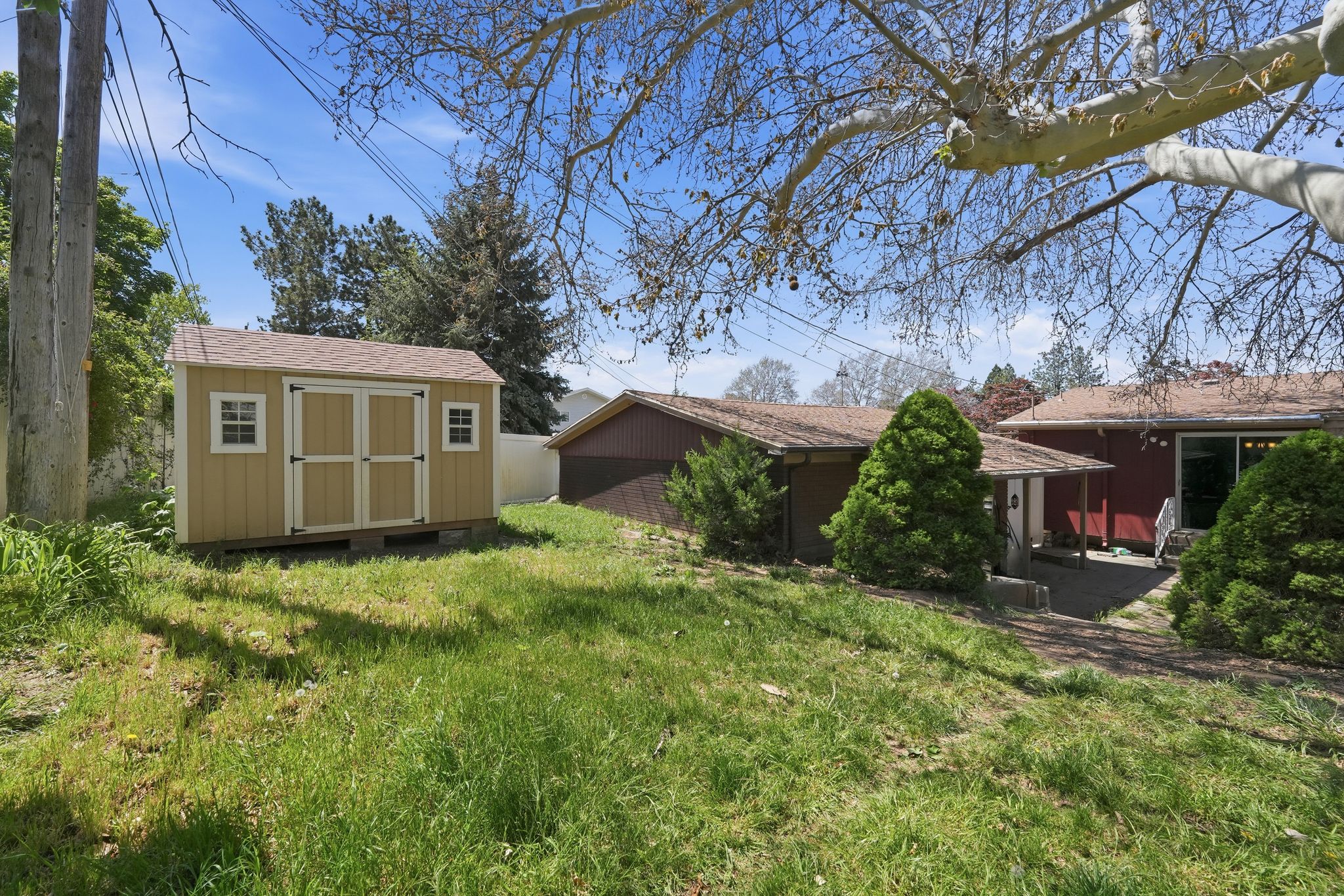 Expansive sloped backyard featuring a freestanding storage shed with double doors and two windows, mature trees, and a visible patio area