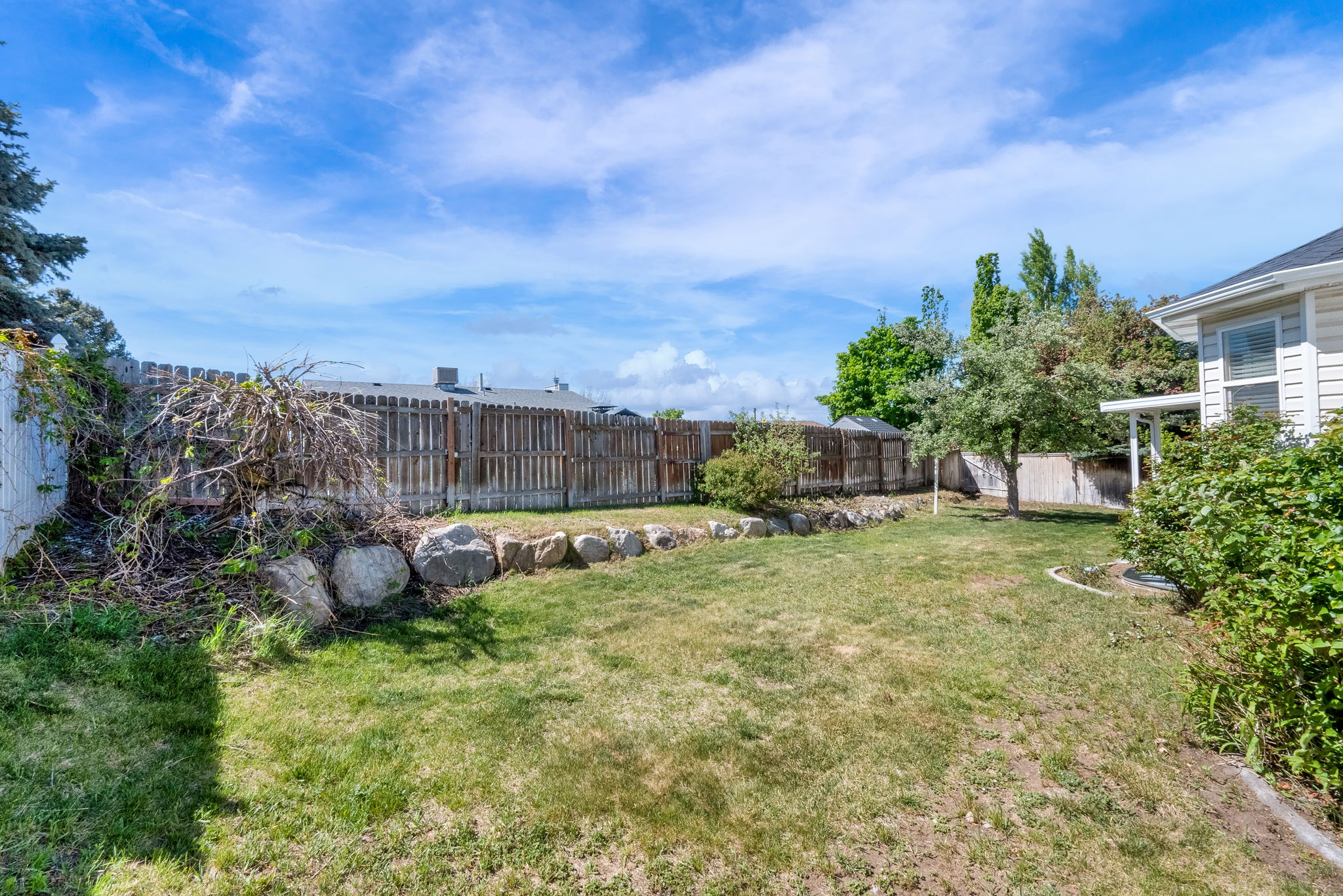 Expansive backyard featuring a manicured lawn, a wood privacy fence, a stone retaining wall, mature trees, and a covered patio area