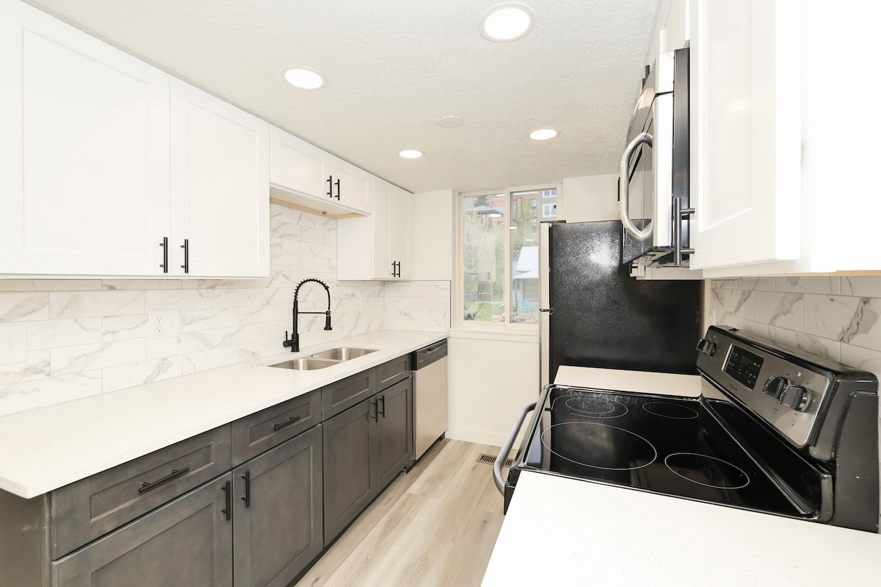 Contemporary kitchen featuring two-tone cabinetry, a dual-basin stainless steel sink with a matte black high-arch faucet, sleek white countertops, and a full tile backsplash