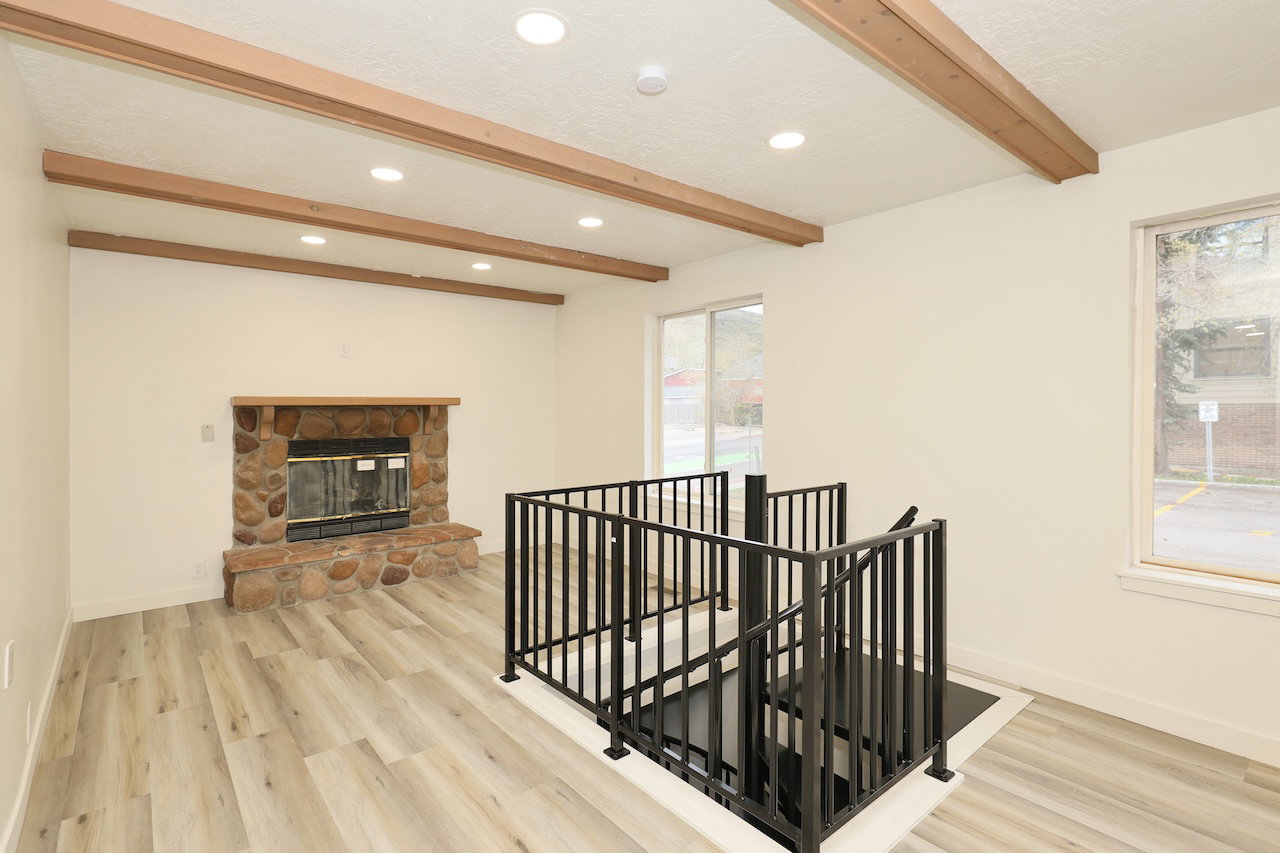 Living area featuring wood-finish flooring, a stone fireplace with a wood mantel, exposed ceiling beams, recessed lighting, and a modern black metal staircase railing