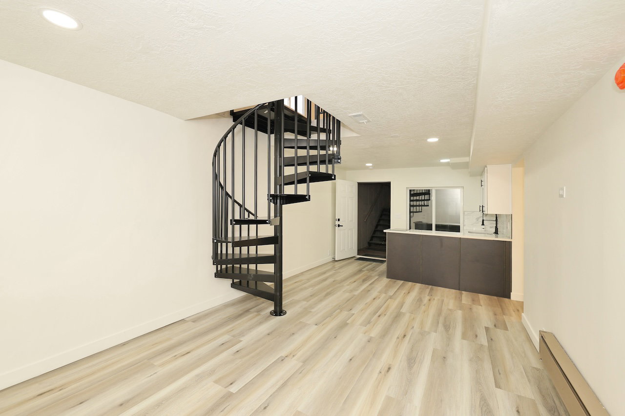 Open-concept living area featuring a black metal spiral staircase, light wood-finish flooring, recessed lighting, white walls, and a built-in counter with white upper cabinetry