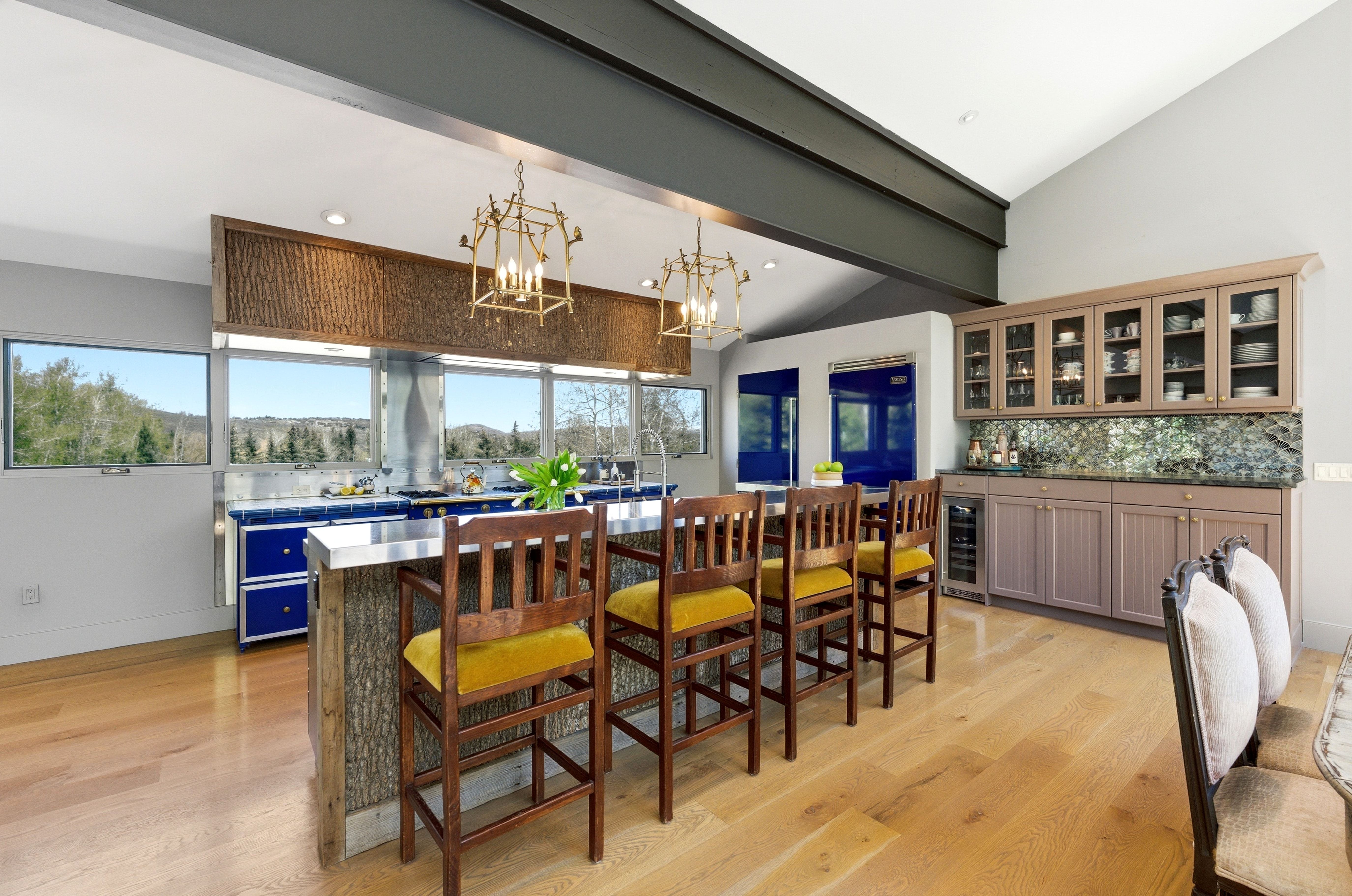 Open-concept kitchen featuring extensive natural light, wood-finish flooring, a wood-clad island with a stainless steel countertop, and a built-in wet bar with glass-front cabinetry