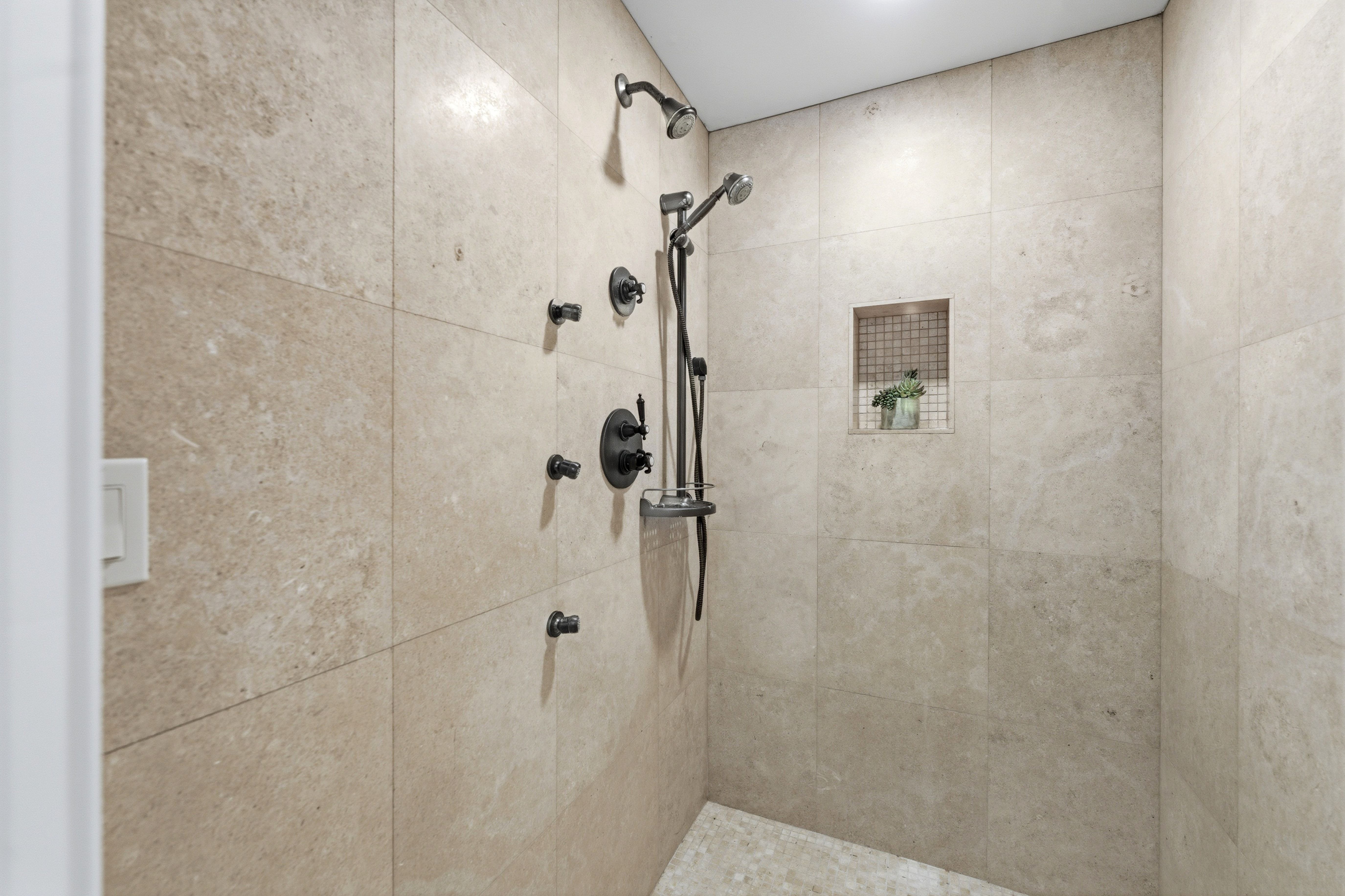 Walk-in shower featuring large format natural stone-look tile, a built-in niche with mosaic tile, a ceiling-mounted rain shower head, a handheld shower fixture, and multiple body jets with a matte black finish