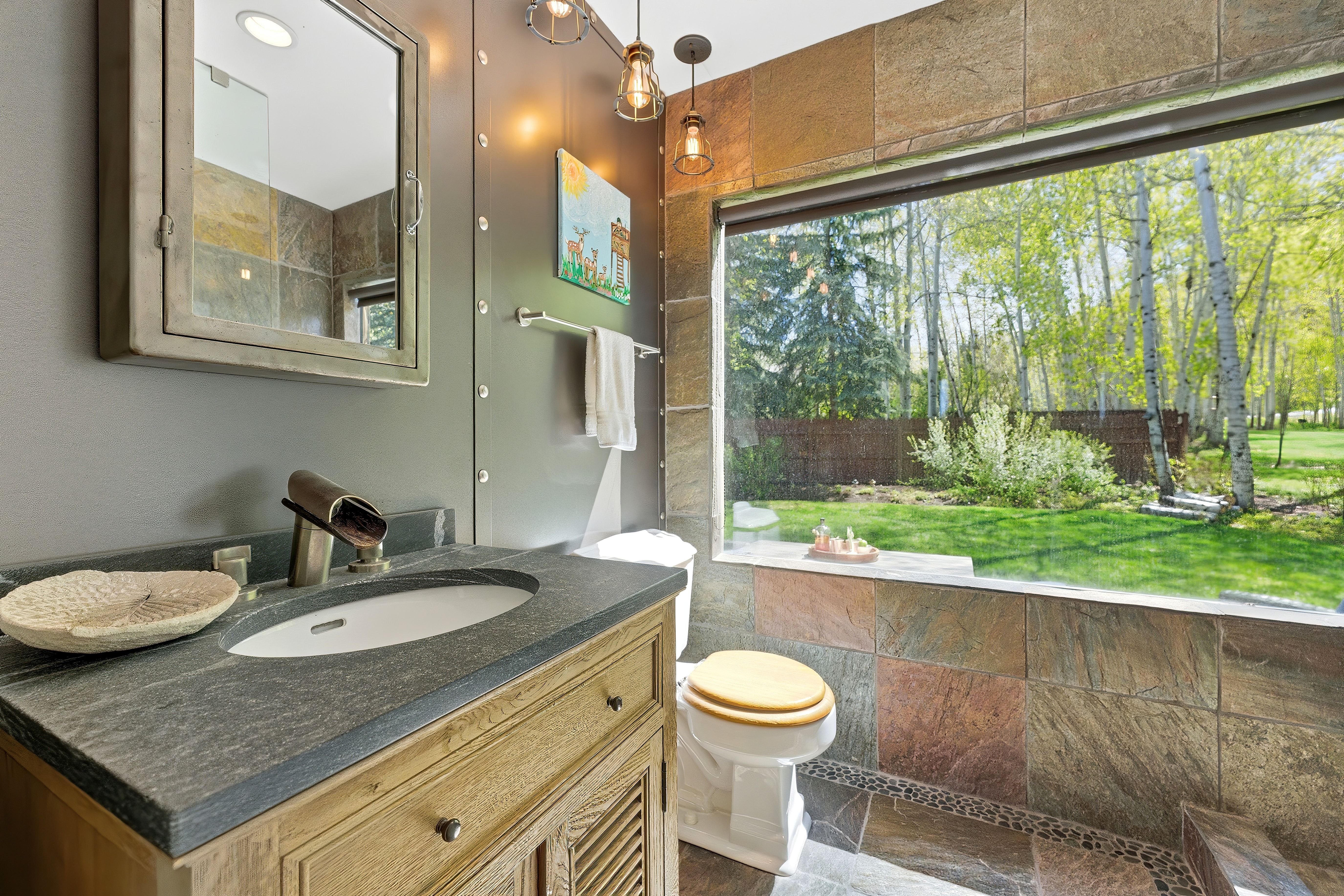 Bathroom featuring an expansive horizontal window, stone tile walls, a stone vanity countertop, a wood-finish vanity cabinet, and industrial-style pendant lighting