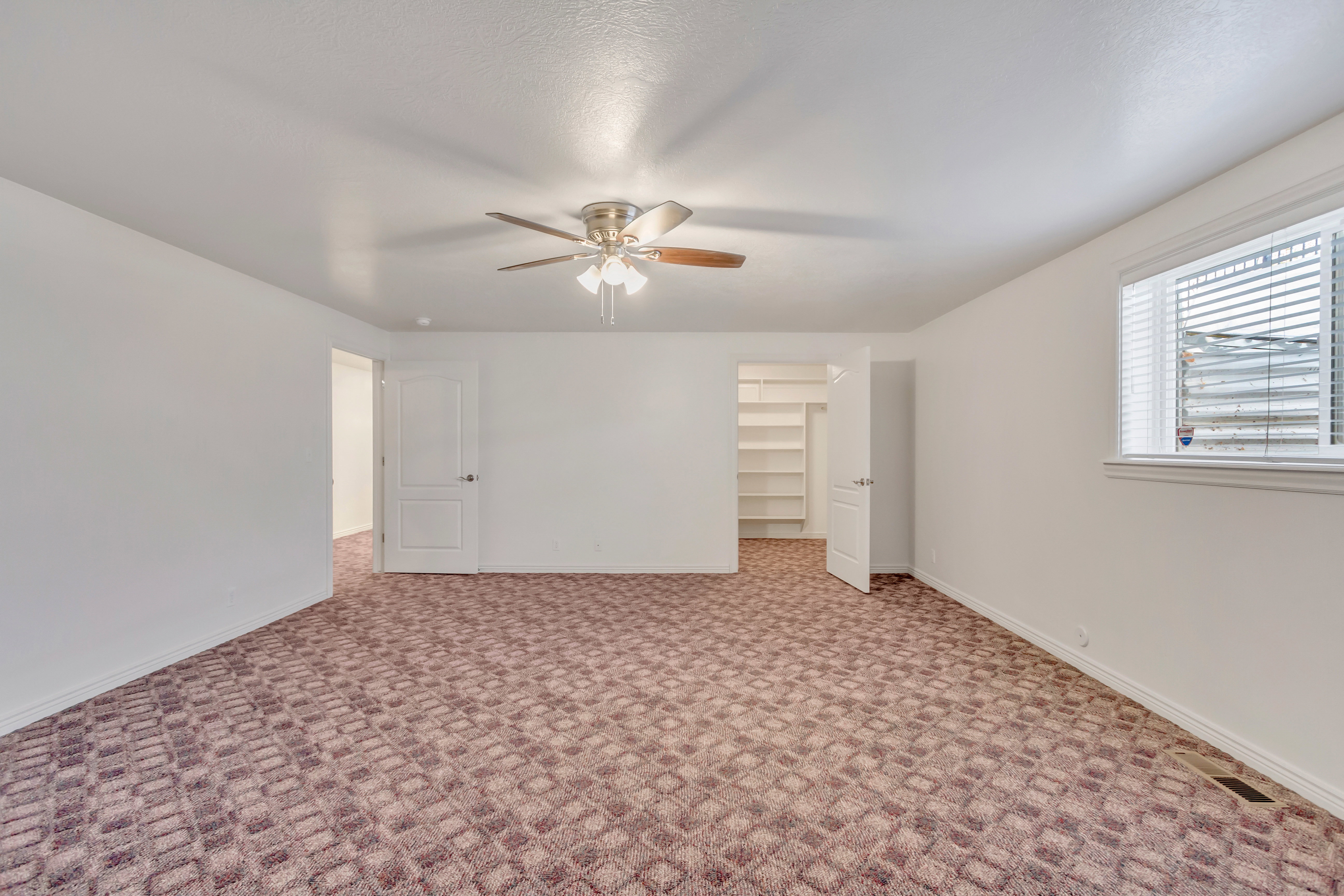 Spacious room featuring patterned carpet flooring, a ceiling fan with light fixture, and a window with horizontal blinds