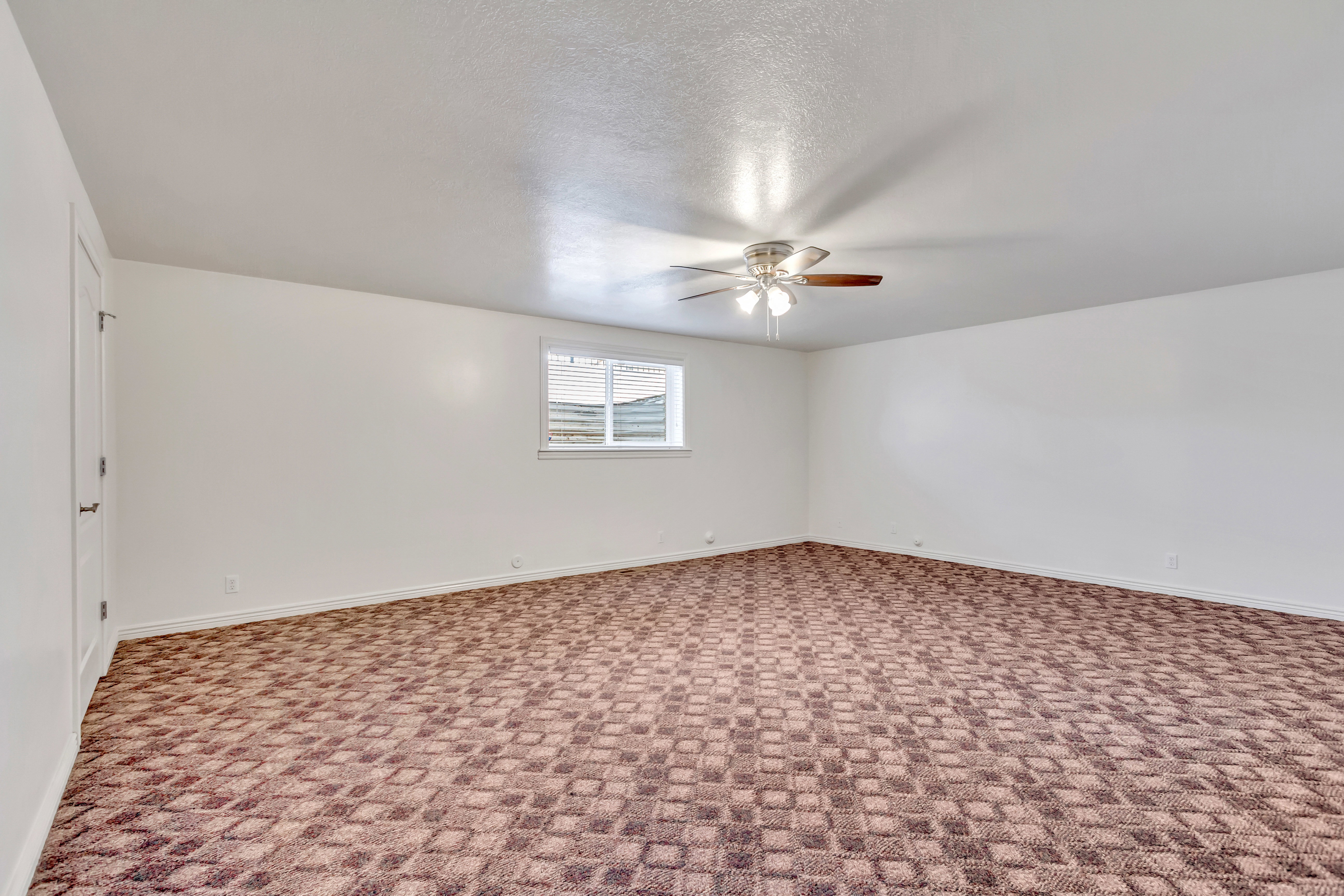 Spacious room featuring patterned carpet flooring, a window providing natural light, and a ceiling fan with integrated lighting