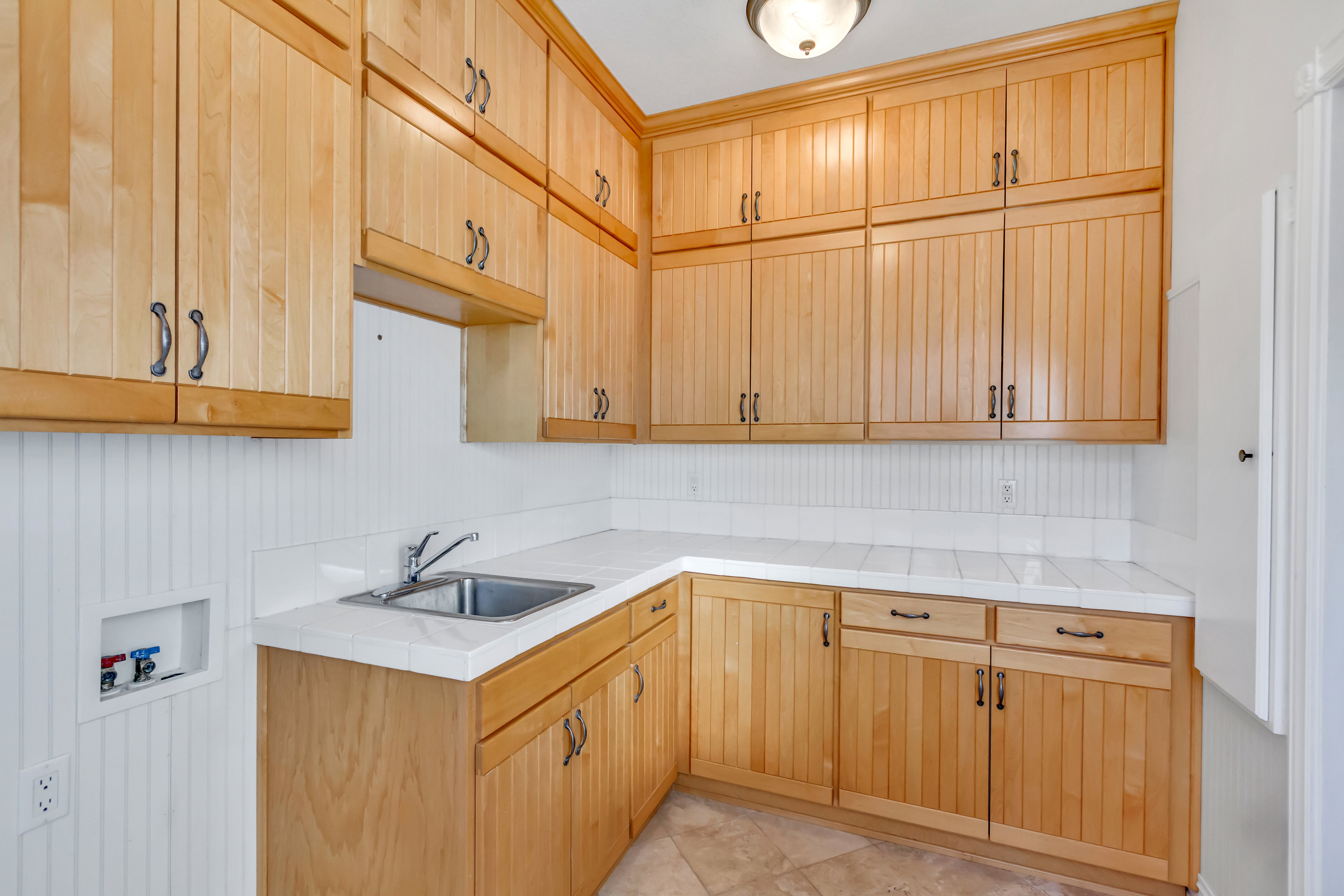 Utility room featuring extensive wood cabinetry, a stainless steel sink, tile countertops, beadboard wainscoting, and tile flooring