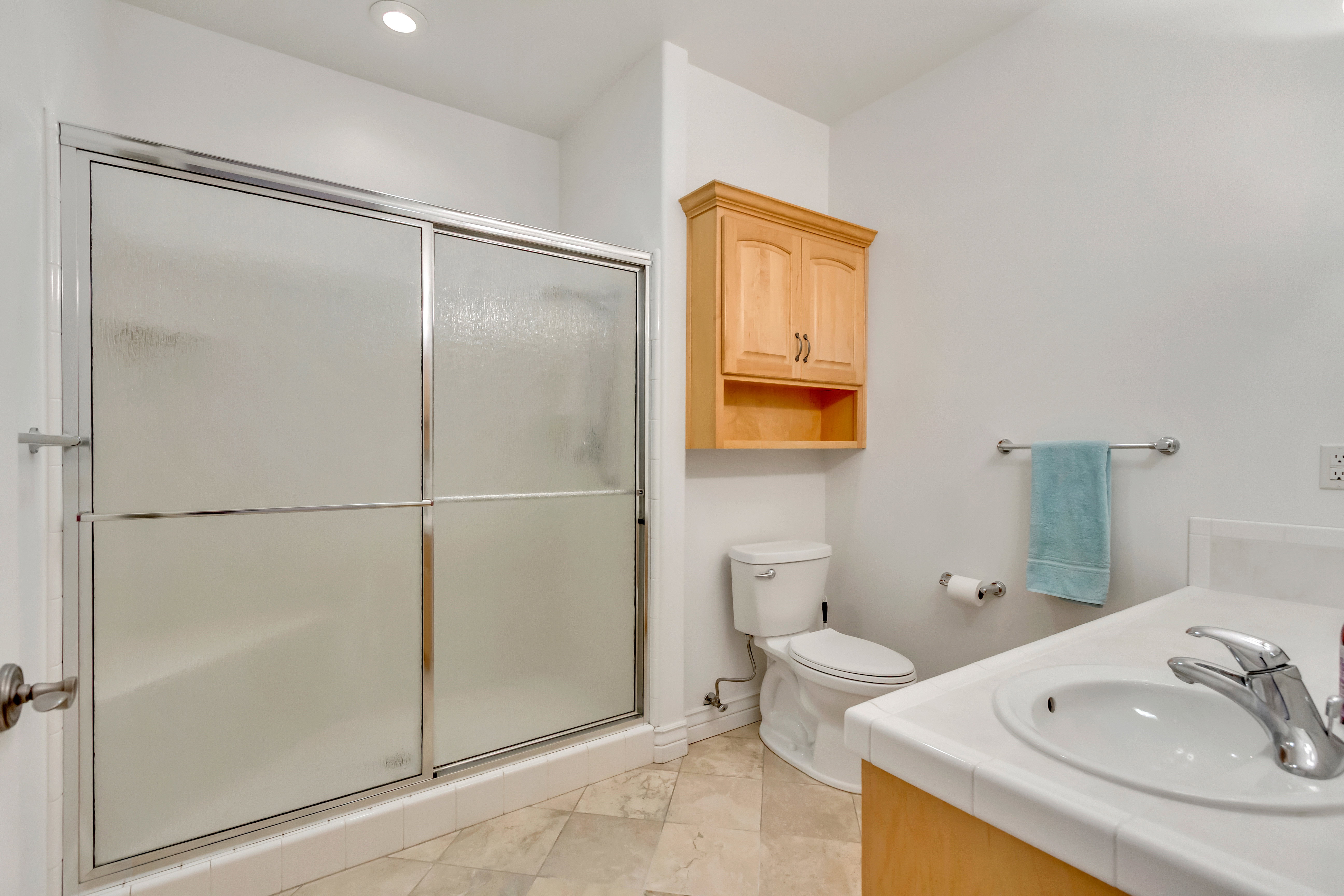 Bathroom featuring a walk-in shower with sliding frosted glass doors, light wood cabinetry, a white countertop sink, and tile flooring