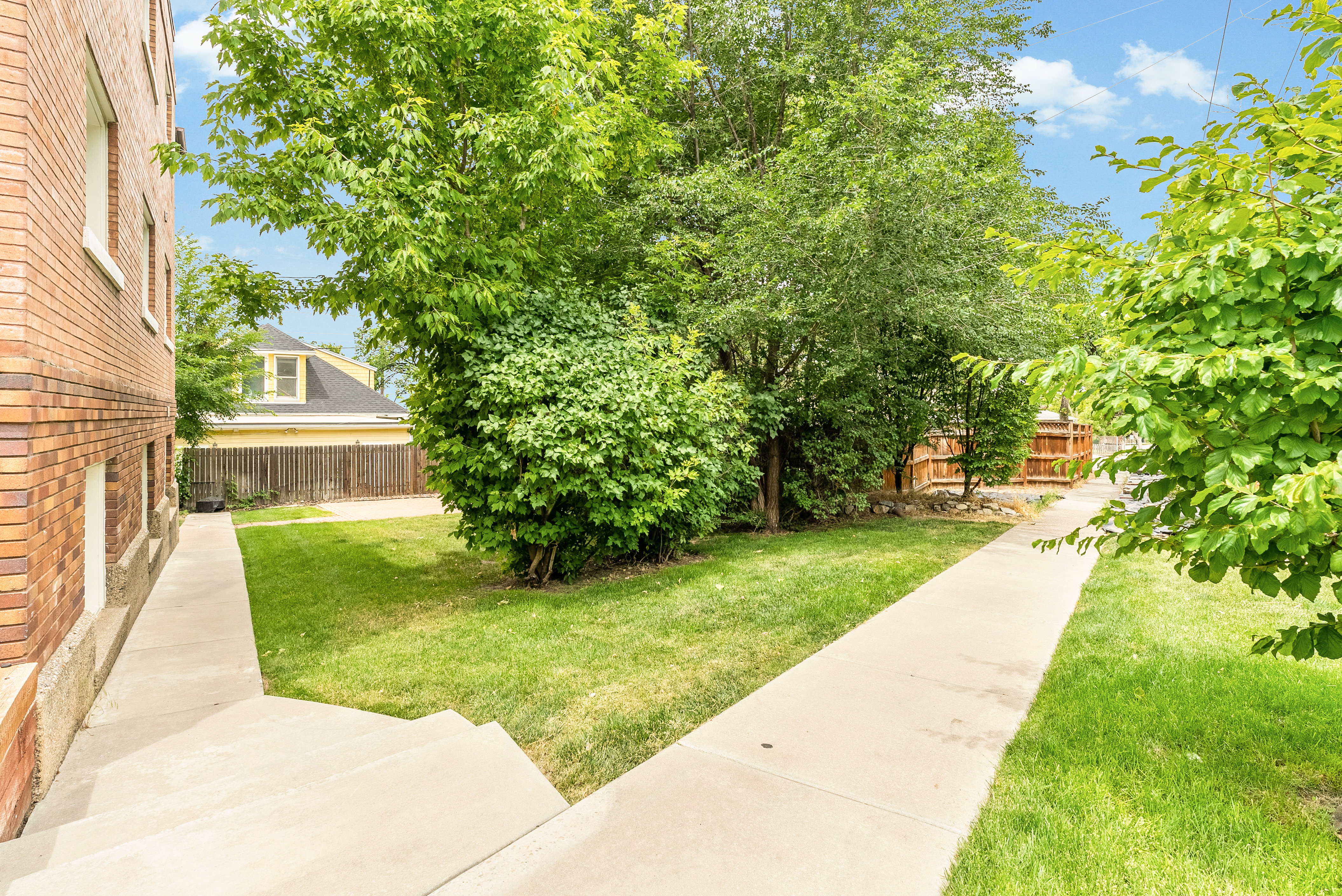 Brick exterior with multiple windows, concrete walkway, and mature trees, complemented by a manicured lawn and wooden fencing