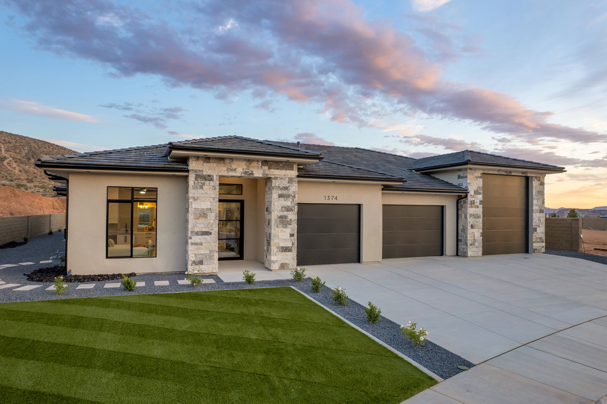 Contemporary stucco exterior with stone accents, featuring a multi-car garage, a flat concrete driveway, an artificial turf lawn, and a desert landscape backdrop