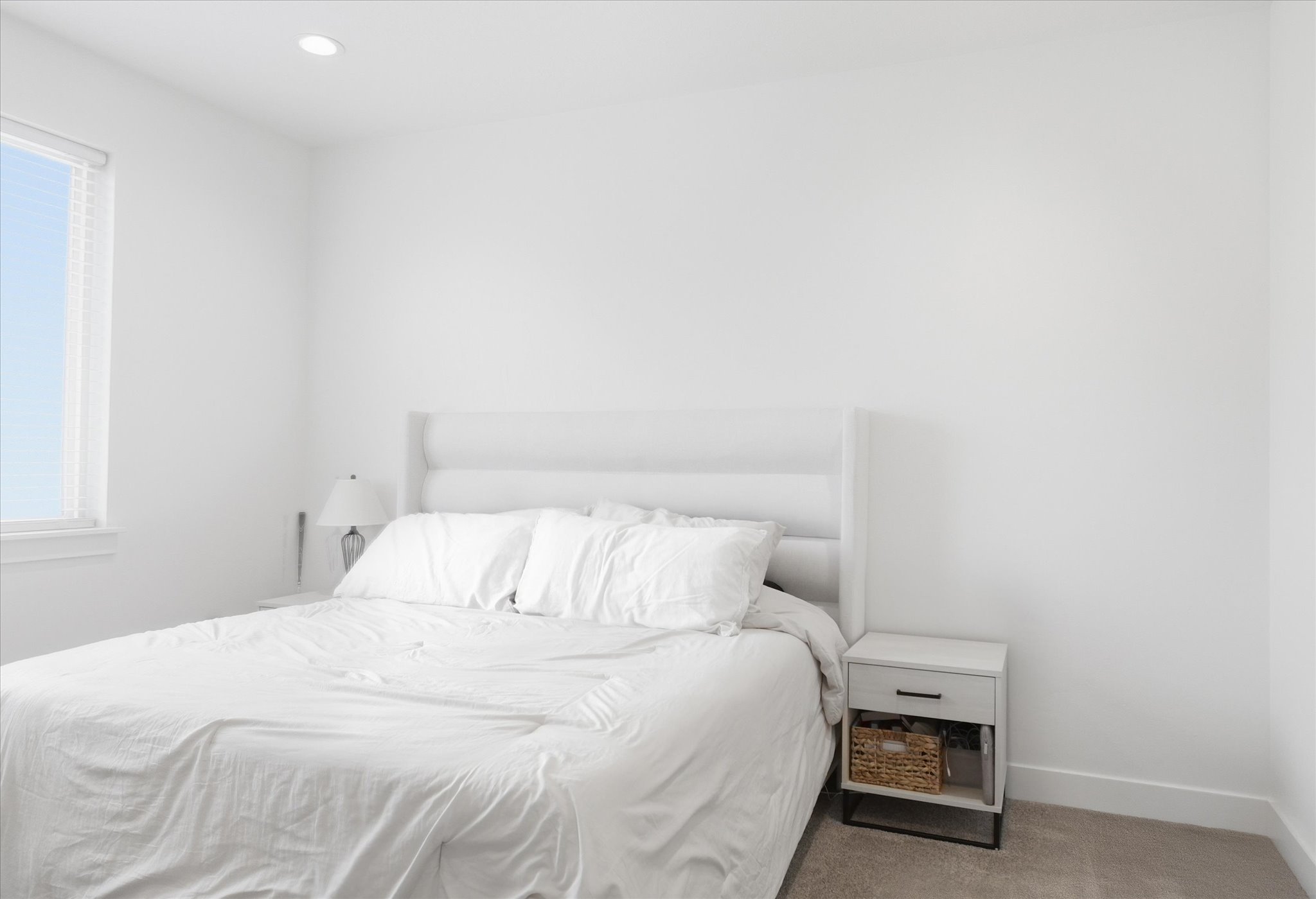 Bright bedroom featuring a recessed light, single window with blinds, light-colored carpet, and a white nightstand with a dark metal frame