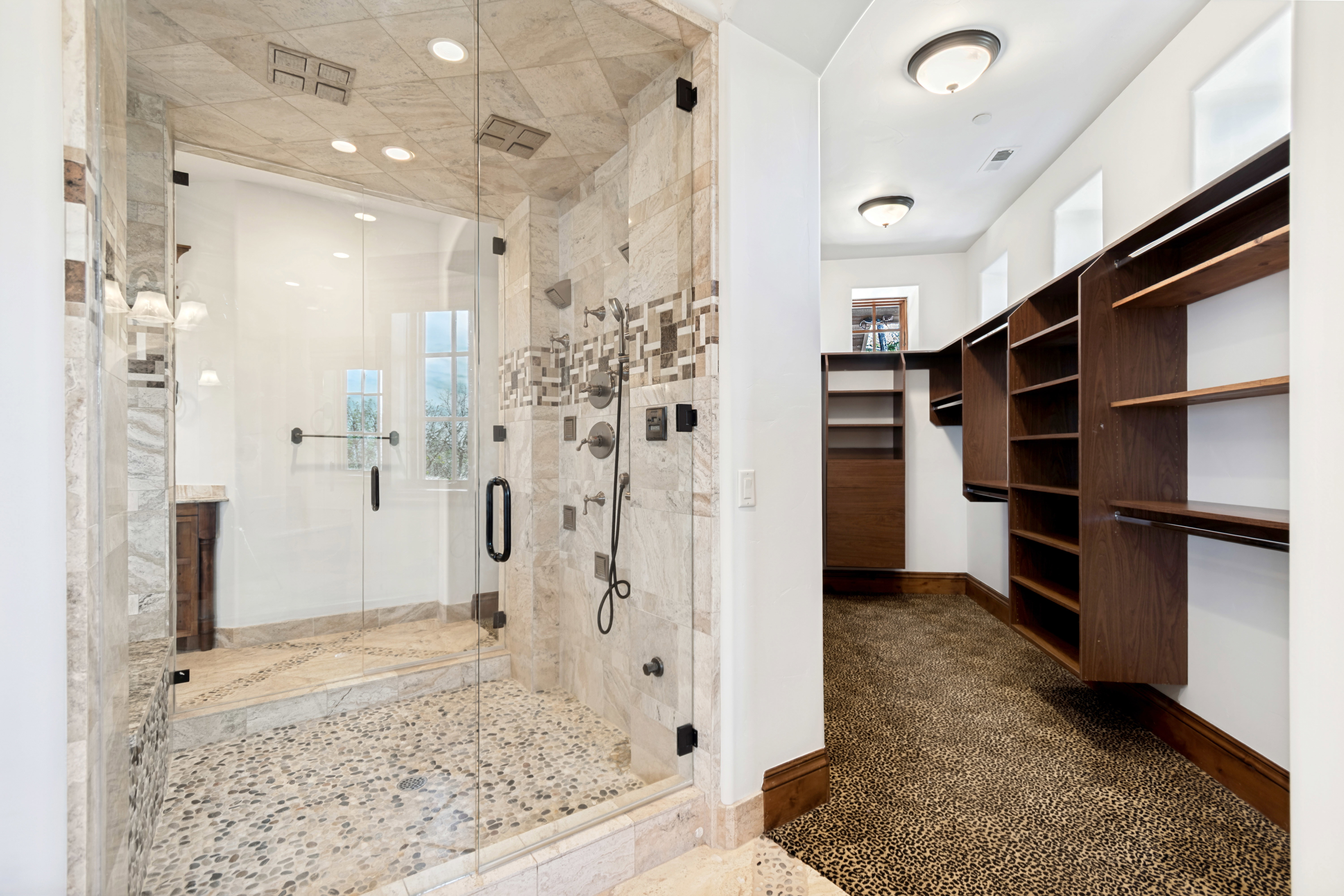 Spacious walk-in shower with river rock flooring, tiled walls, and a multi-function shower system, complemented by an adjacent closet featuring custom wood-finish shelving and textured carpeting