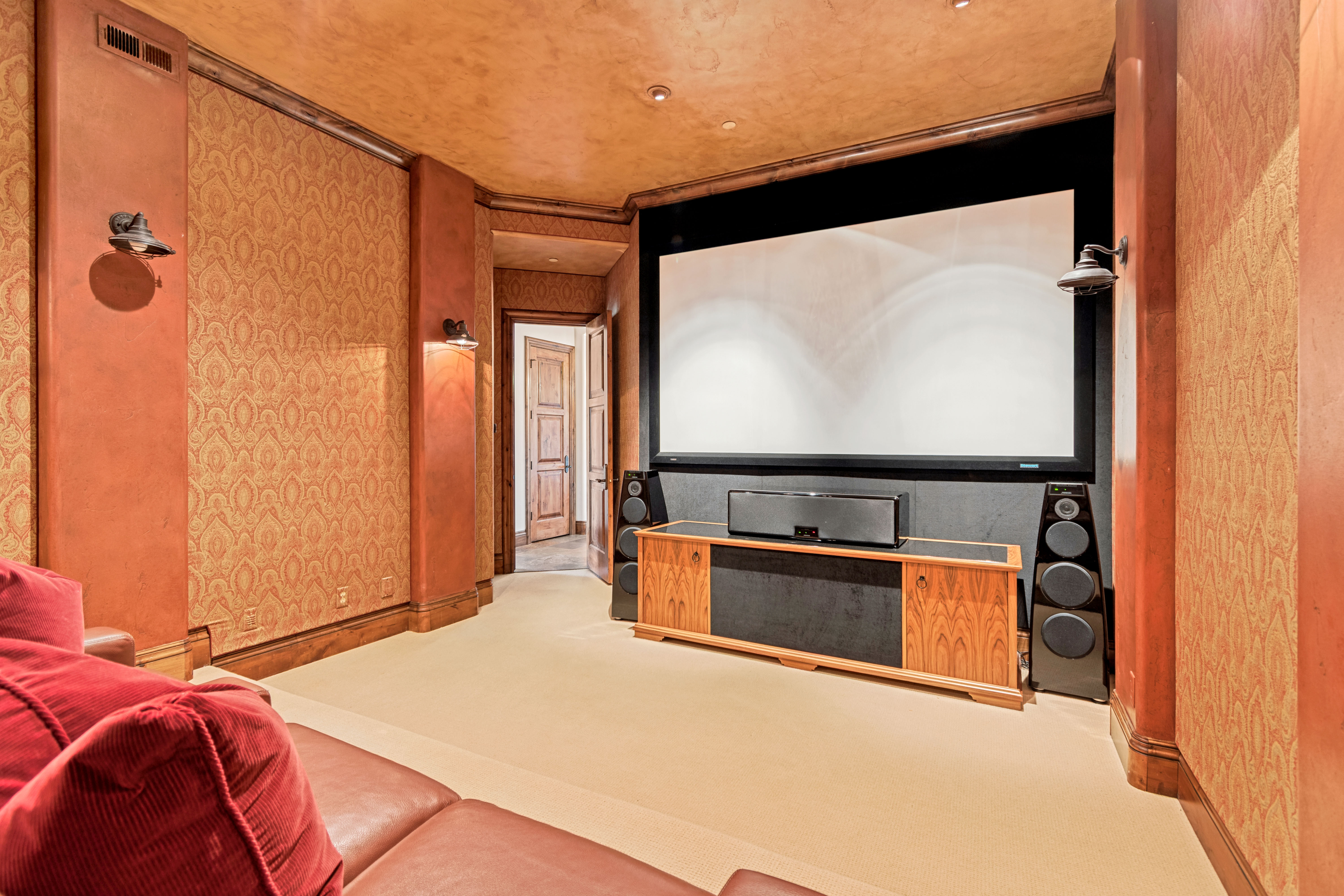 Dedicated media room featuring a large projector screen, built-in speaker system, wall sconces, patterned wall coverings, and an adjacent wood door